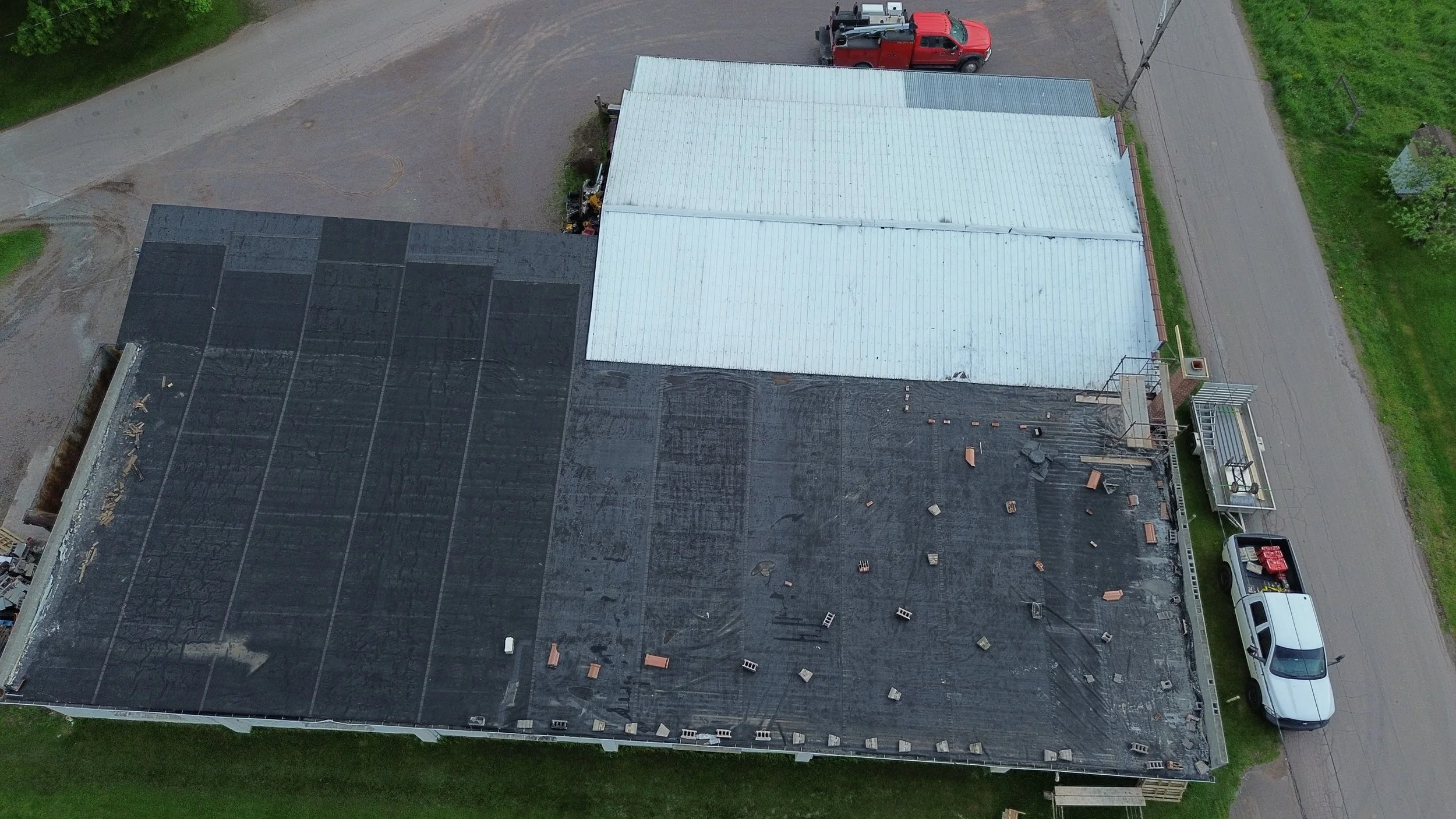 An aerial view of a building with two sections, one with a black roof and the other with a white roof, undergoing roofing work. Construction materials and debris are scattered on the black roof. A red truck and a white van are parked nearby, with a dirt road and green grass surrounding the area.