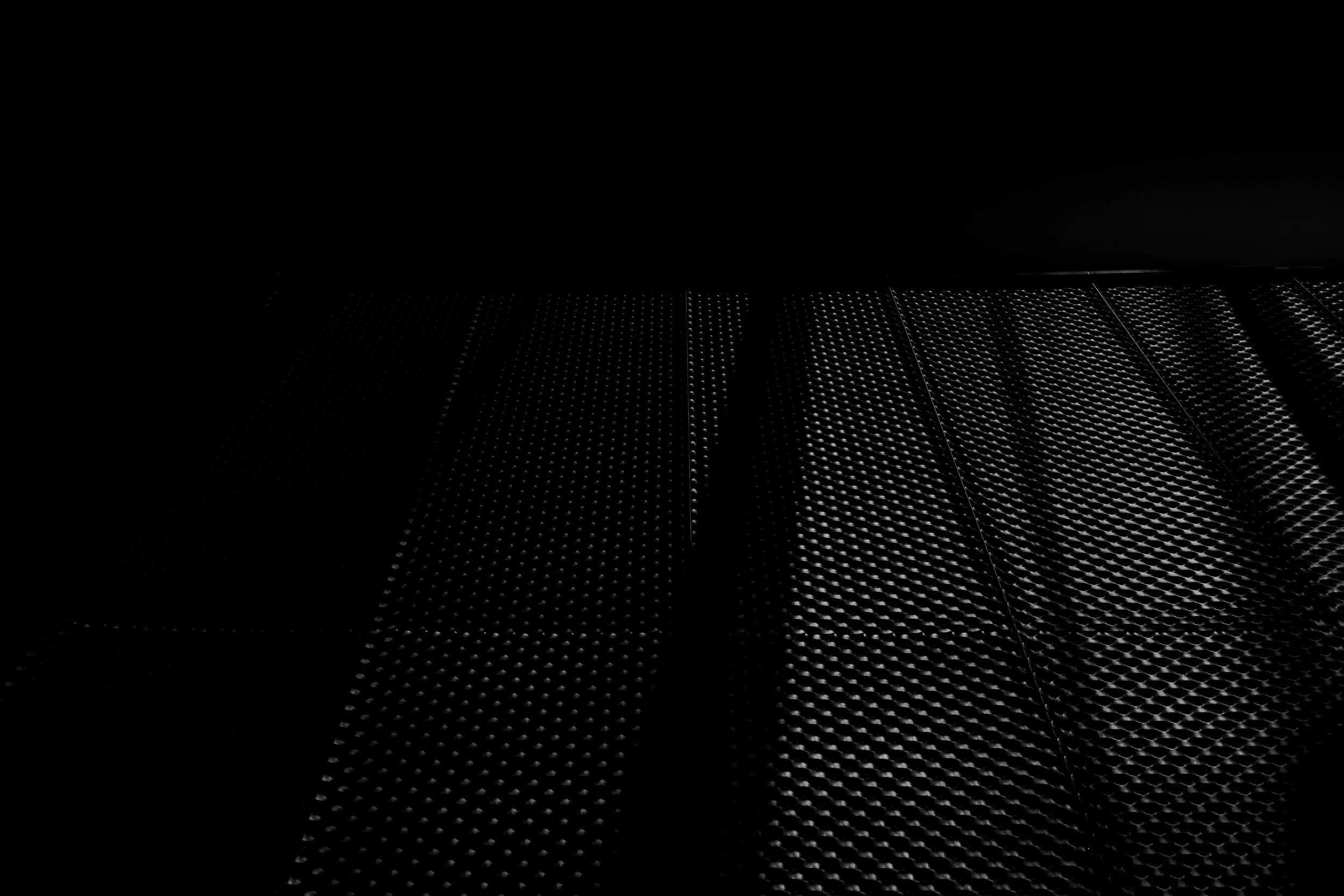 Close-up of a dark metallic surface with a textured, dot pattern, possibly part of industrial equipment or furniture, illuminated by light from the right side, creating shadows on the left.