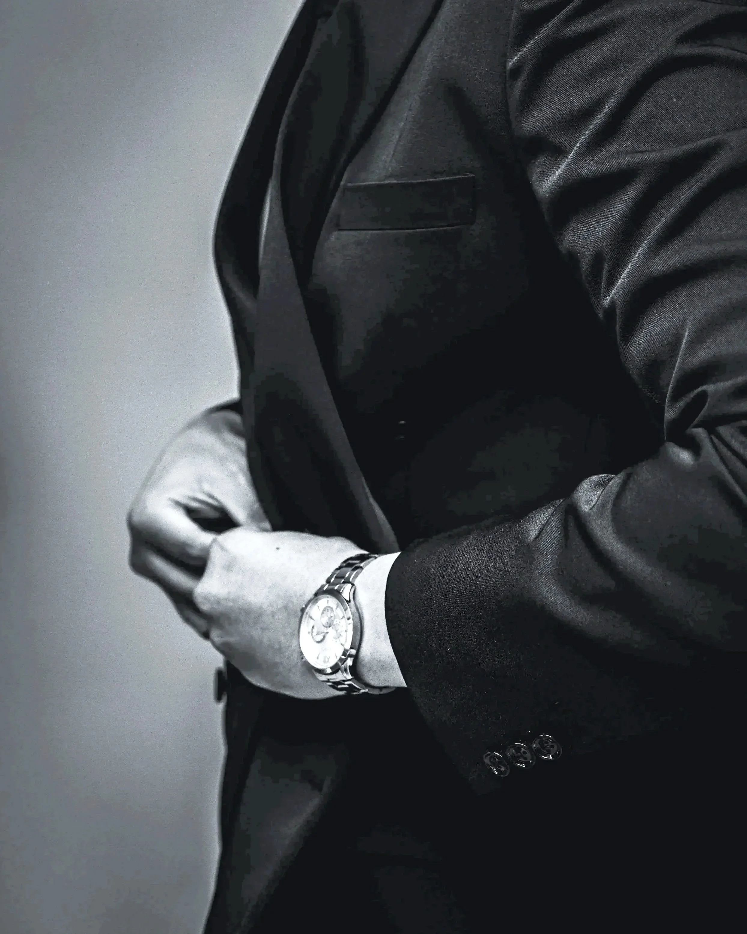 A grayscale / black-and-white close-up image of a person buttoning up his suit jacket, possibly an attorney or a lawyer, dressed in a black suit, wearing a silver wristwatch, standing against a gray background.