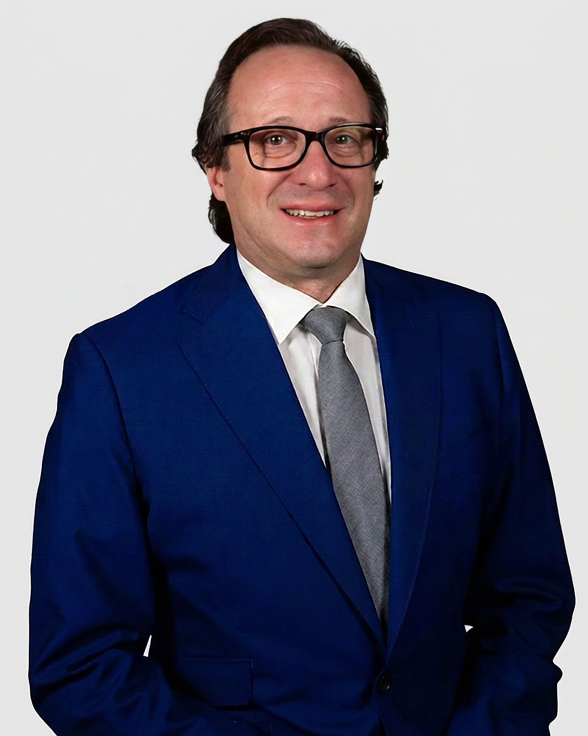 A professional headshot of a man with glasses and dark hair smiling, dressed in a dark blue suit, white shirt, and gray tie, against a white background.