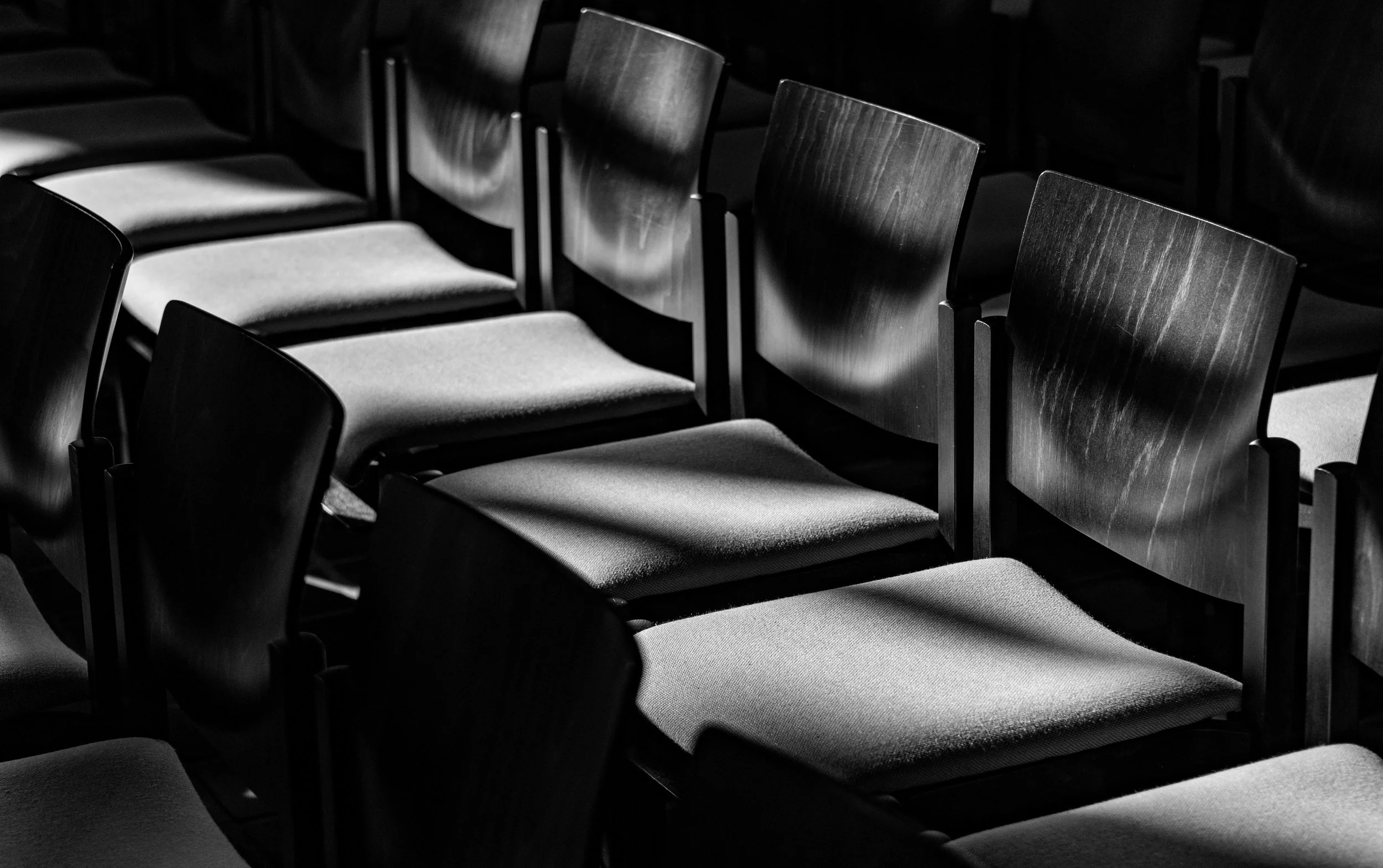 A grayscale / black-and-white image of a row of wooden chairs with fabric seats arranged in a dimly lit area, with shadows cast on the chairs, possibly in a court room.