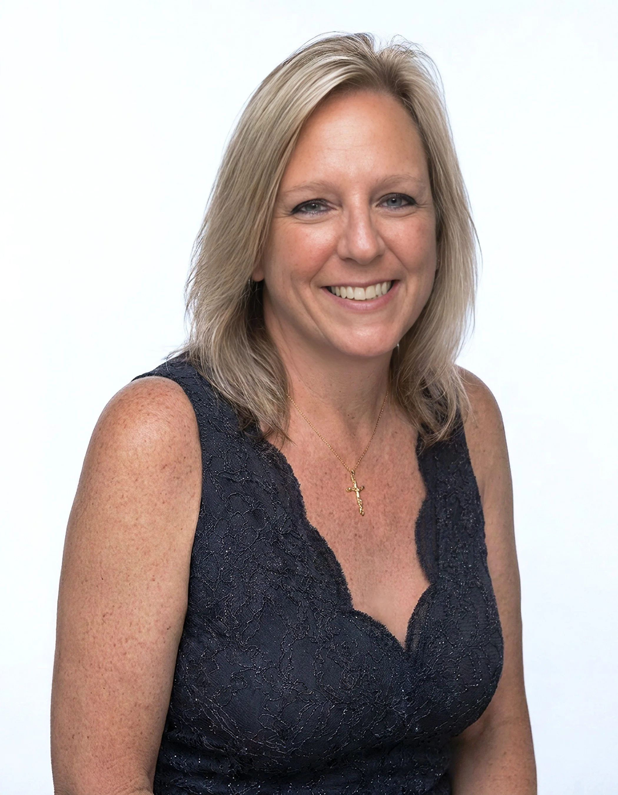 A professional headshot of a woman with blonde hair smiling, wearing a navy lace sleeveless top, a gold cross necklace, and standing against a white background.