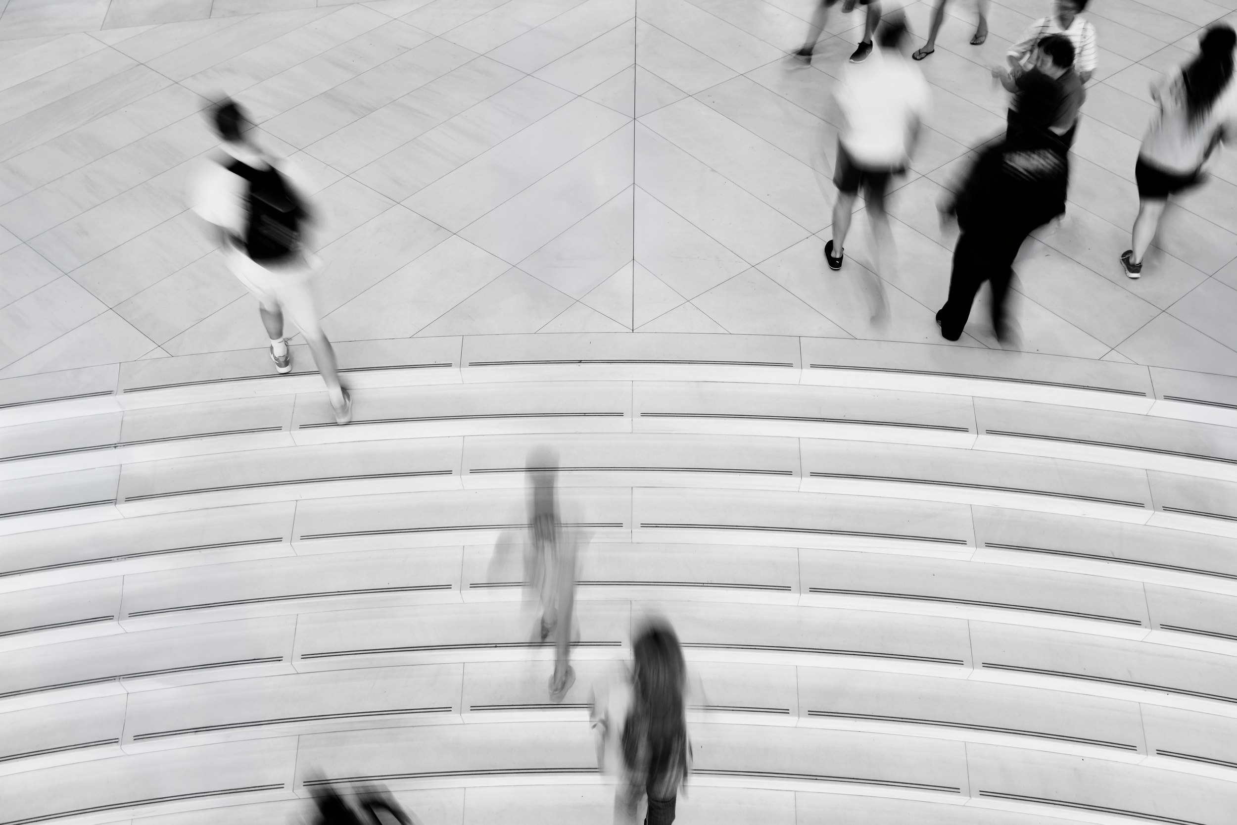 A grayscale / black-and-white image of people walking up or standing near the stairs in a court building or public indoor space, captured from a high angle, with some people blurred due to motion.