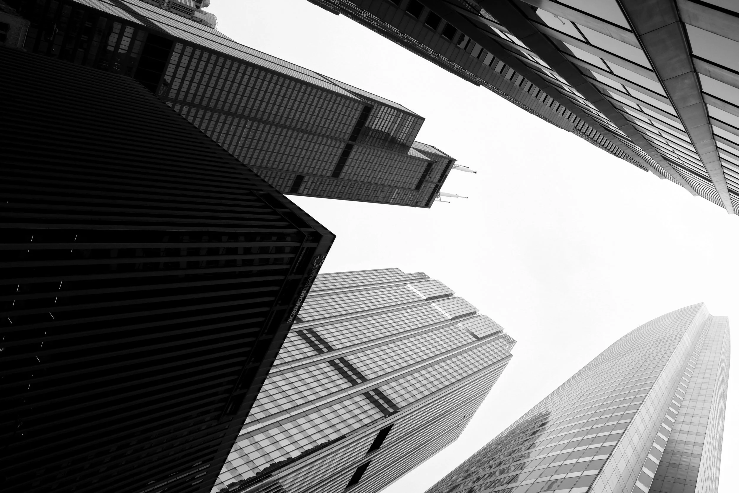 A grayscale / black-and-white photograph taken from the ground looking up at tall skyscrapers in a city of Chicago, with modern glass buildings and architectural details reaching towards the sky.