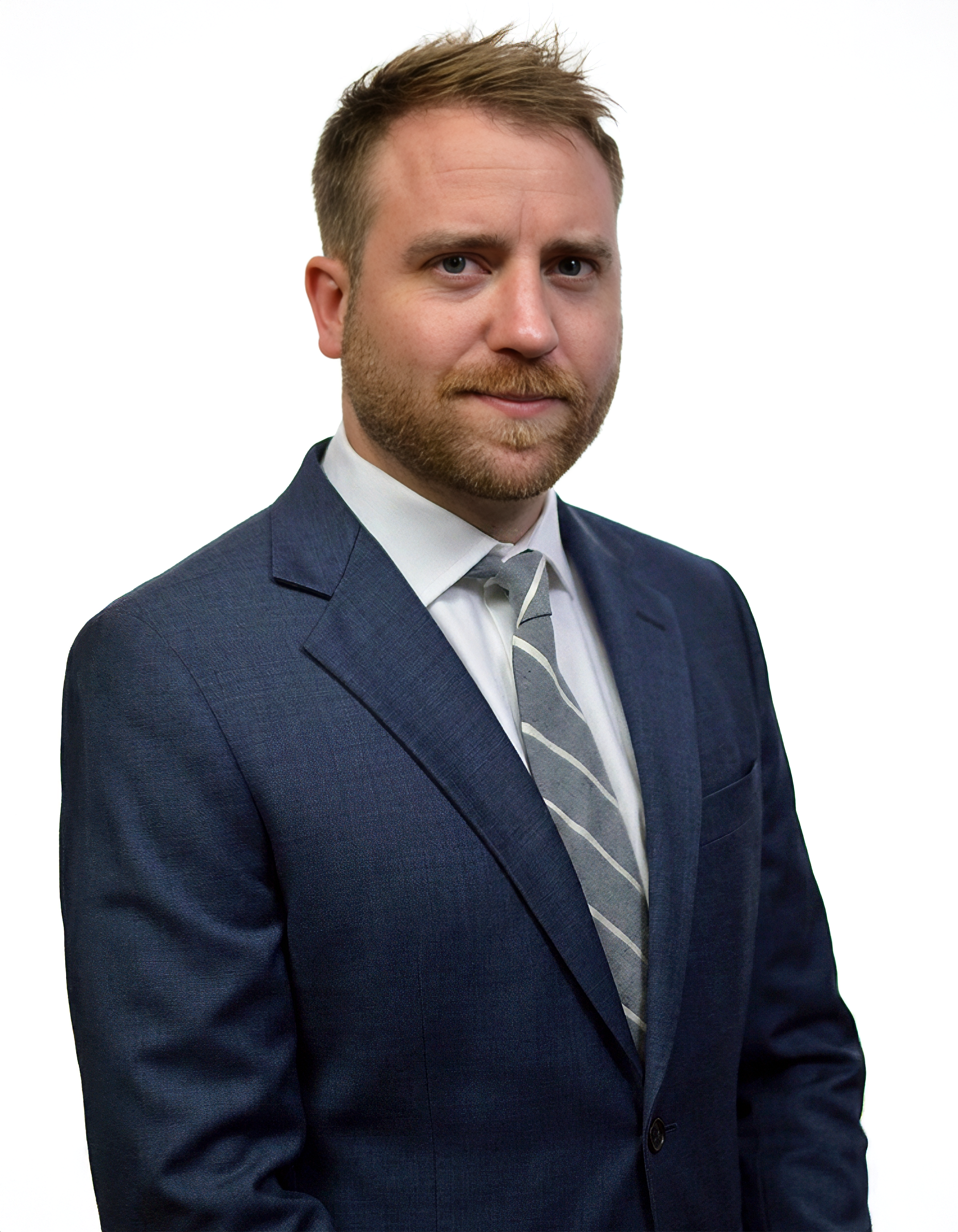 A professional headshot of a man with light brown hair, a beard, wearing a navy blue suit, light blue dress shirt, and a striped tie, against a white background.