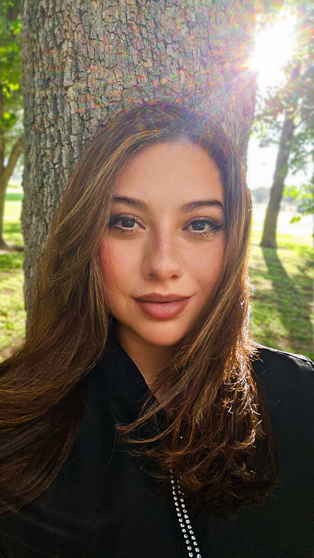 A woman with long brown hair smiling outdoors in front of a tree with sunlight shining through.