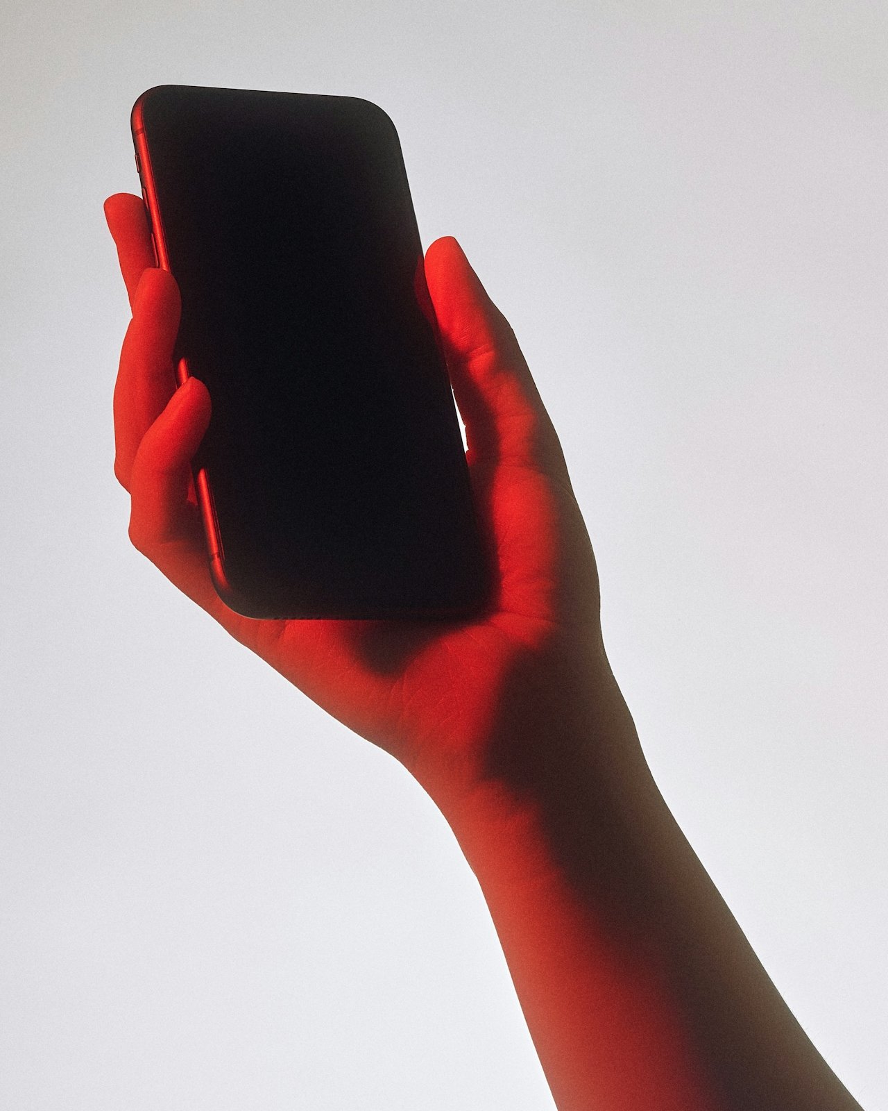 A hand holding a smartphone with a black screen, illuminated by red light.