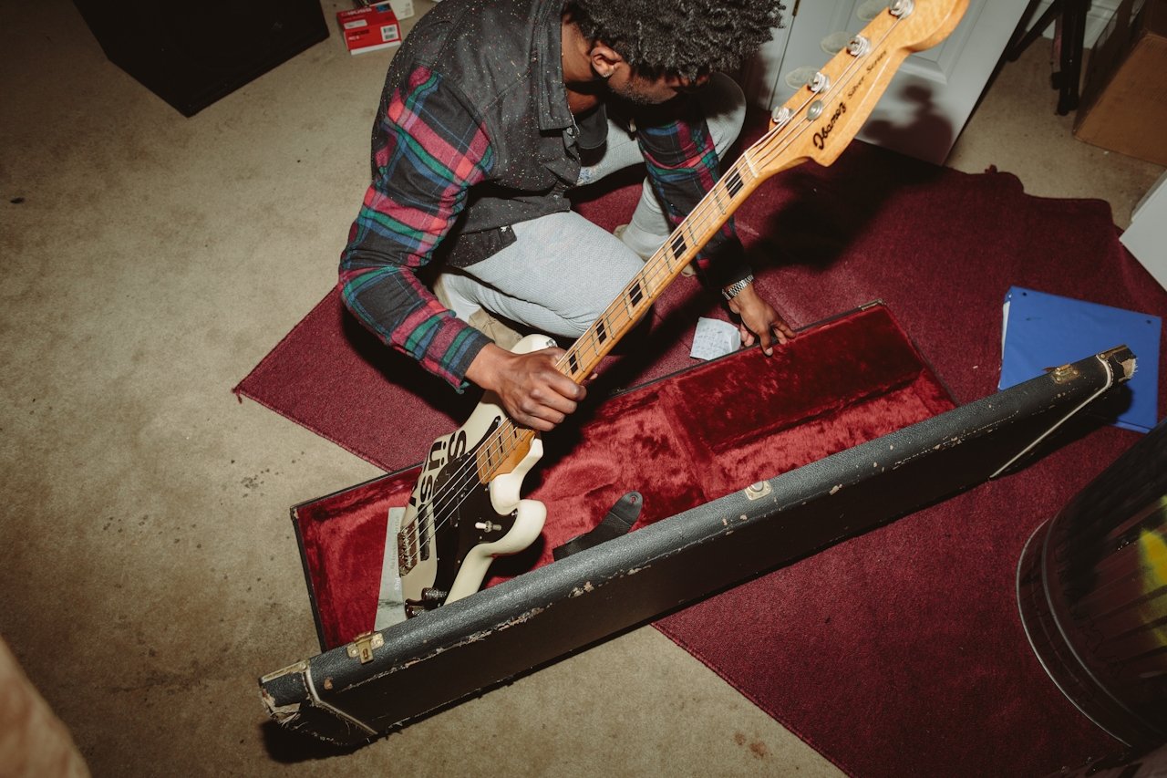 A person with curly hair and glasses wearing a plaid shirt and light gray sweatpants is kneeling on a red carpet, opening a guitar case with a white bass guitar inside. The scene is set in a room with a beige carpet, cardboard boxes, and a trash can nearby.