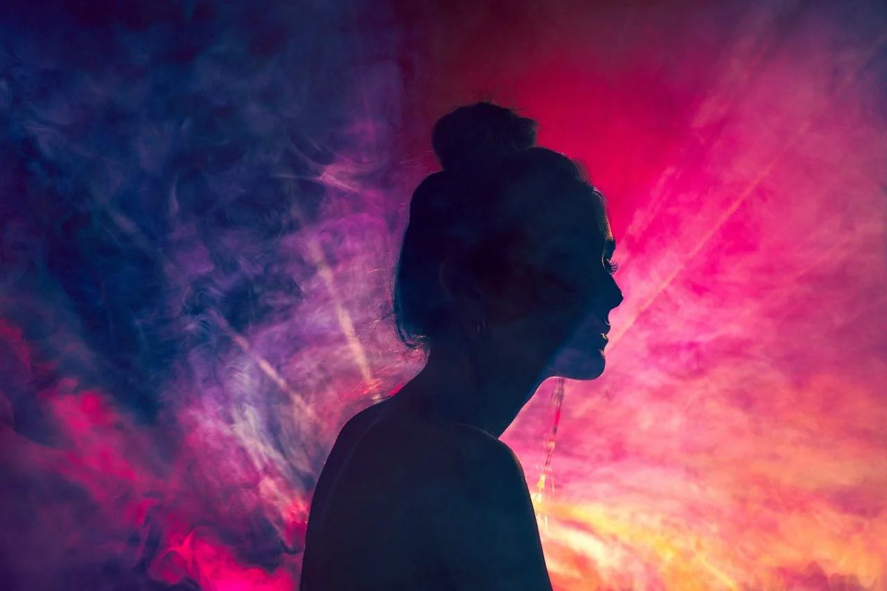 Silhouette of a woman with a bun hairstyle against a colorful, smoky background with pink, purple, and orange hues.