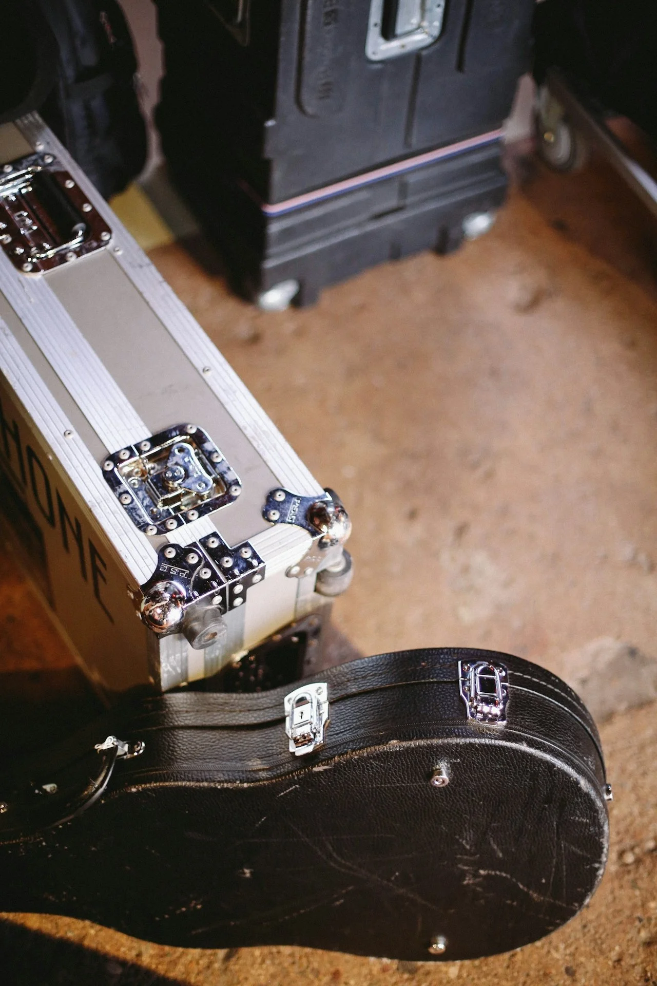 Close-up of a black musical instrument case next to a silver and brown hard case, with other cases partially visible in the background on a brown surface.