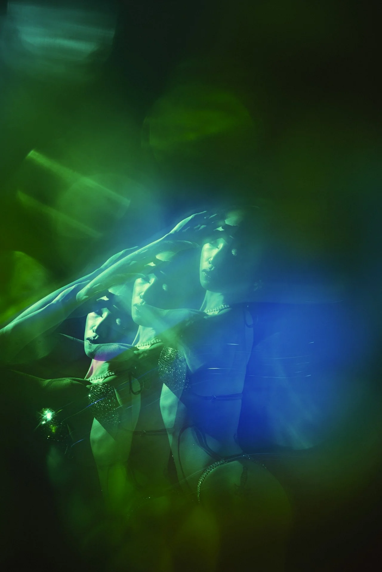 A person posing with their hand near their face under green and blue neon lights, wearing a shiny, rhinestone-studded outfit.