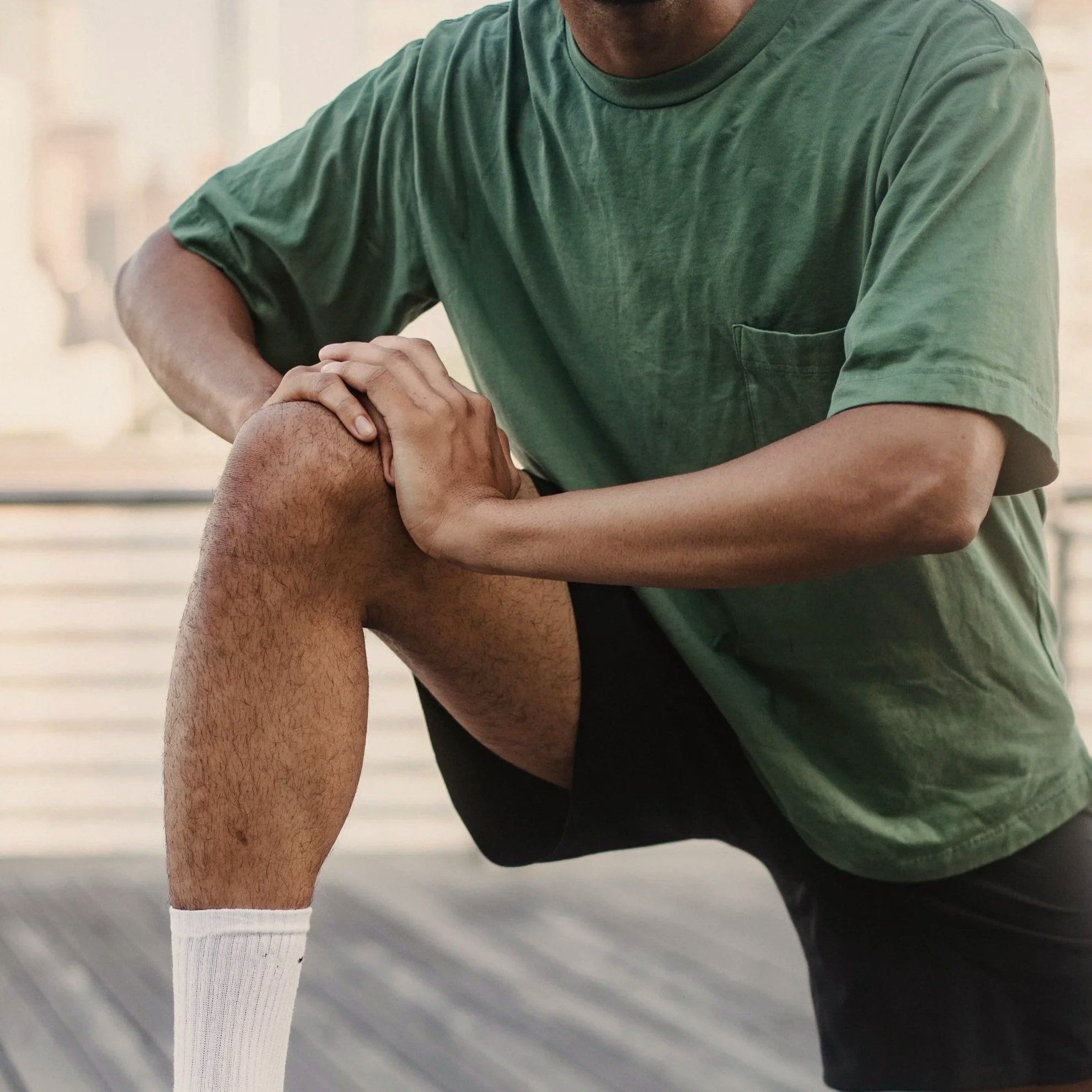 A person in a green t-shirt and white socks is bending forward, holding their sore knee with both hands.
