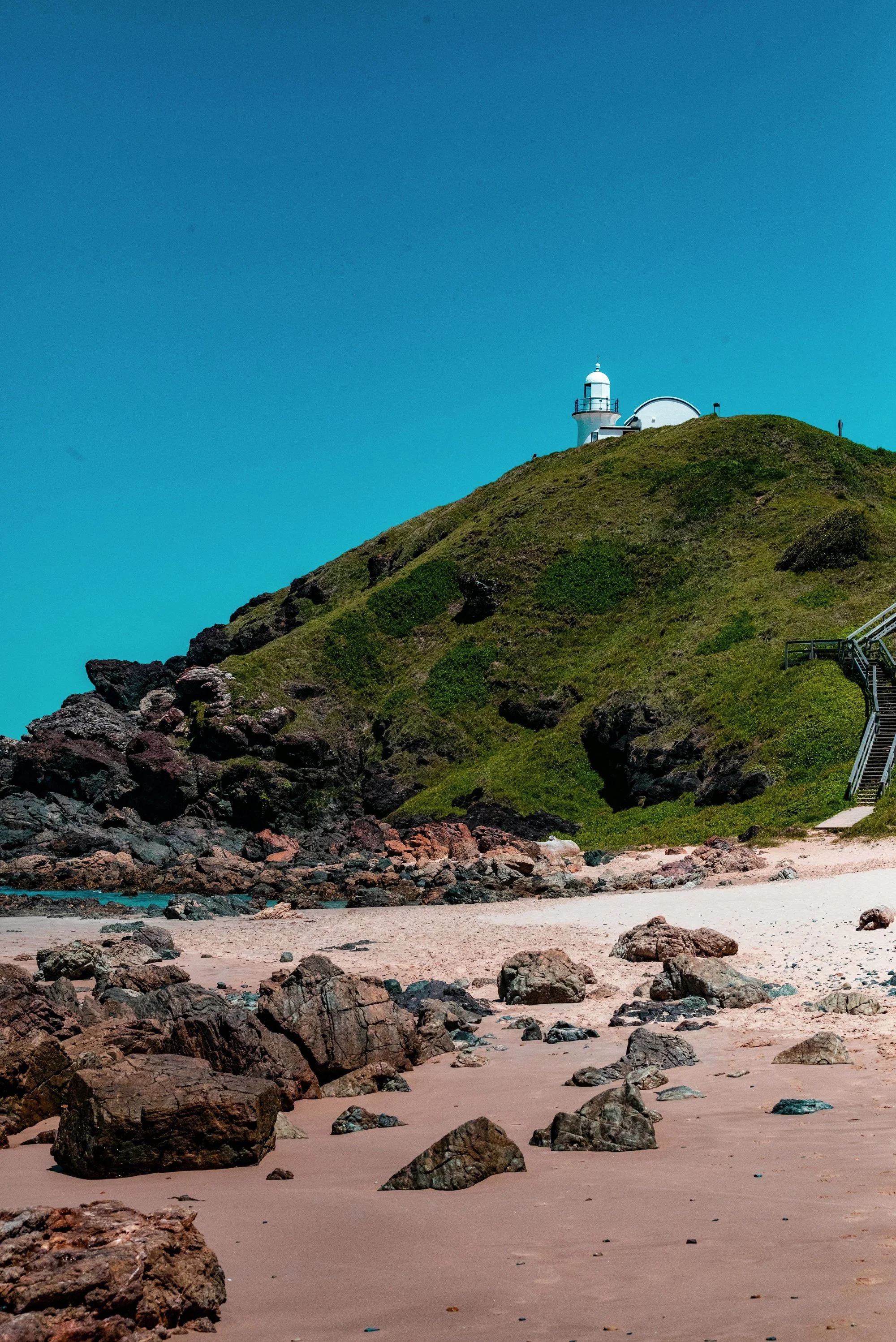 View of a lighthouse on top of a grassy hill near the ocean, with rocks and sand in the foreground and a clear blue sky.