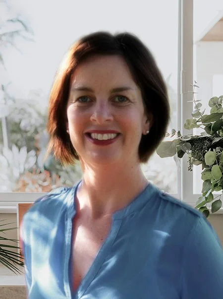 A woman with short dark hair wearing a light blue shirt standing near a window with greenery in the background.