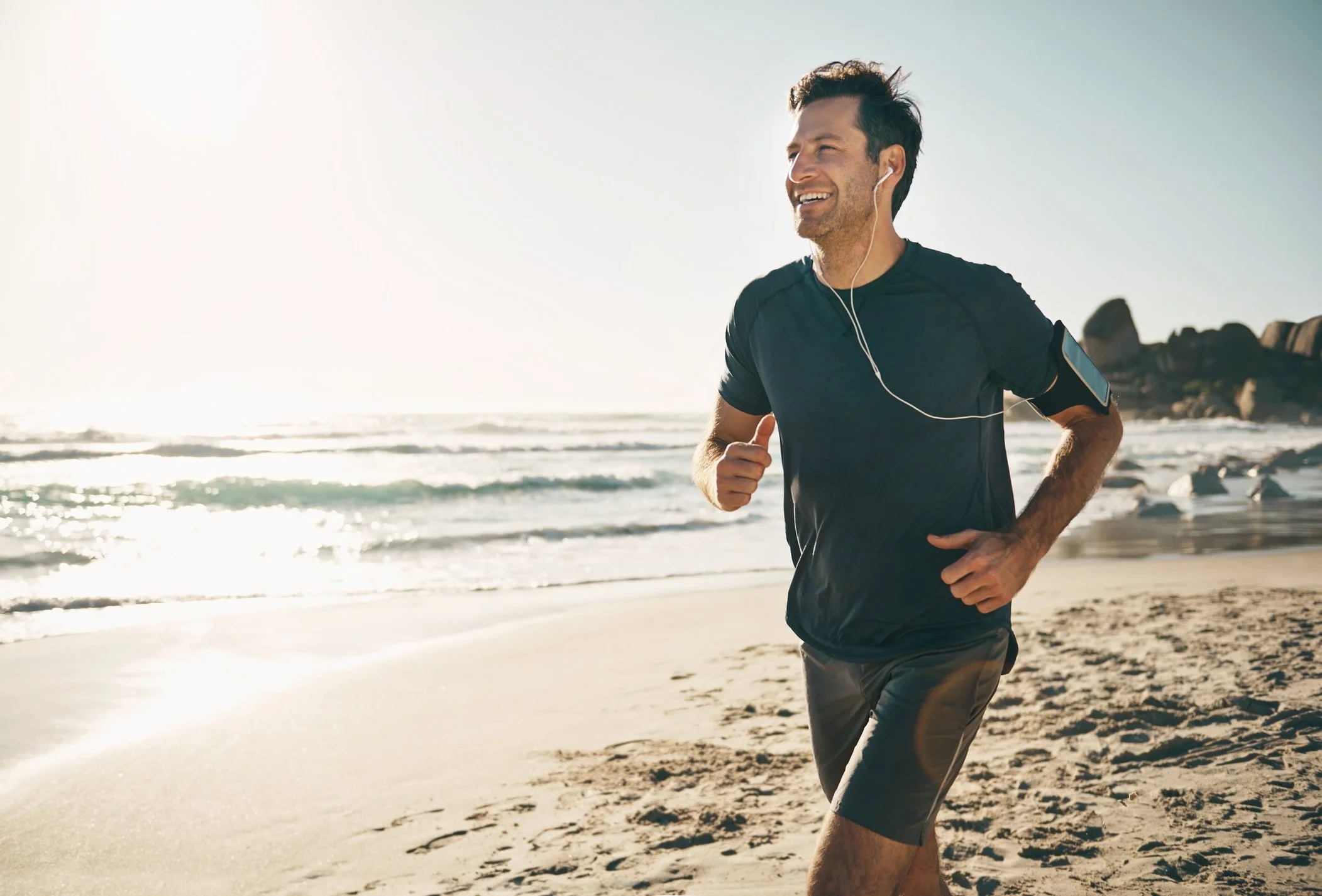 A man running on the beach, smiling, wearing earphones, a black athletic t-shirt, and shorts during sunrise or sunset.