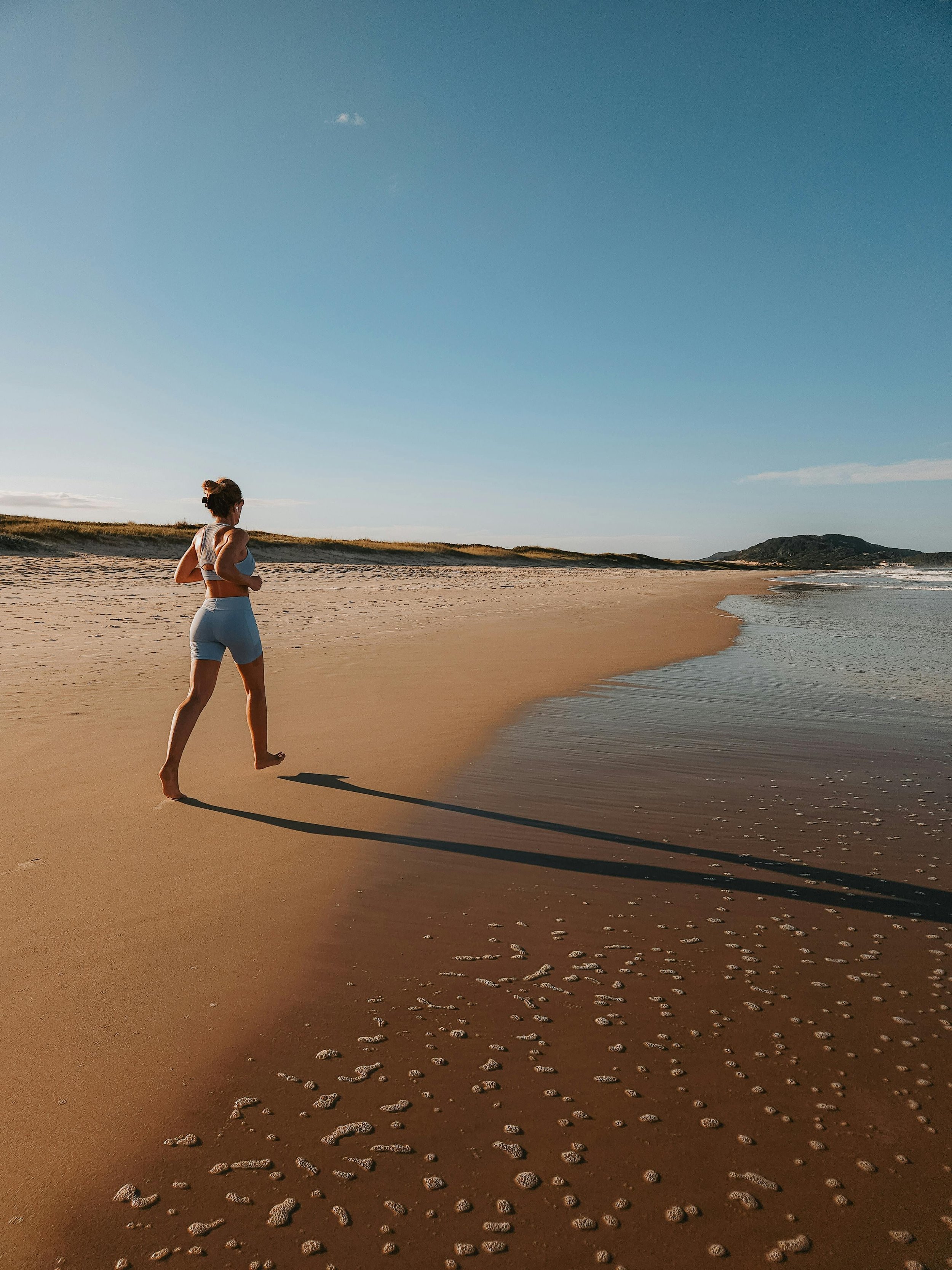 A woman jogging on a sandy beach during sunset with footprints behind her and distant hills.