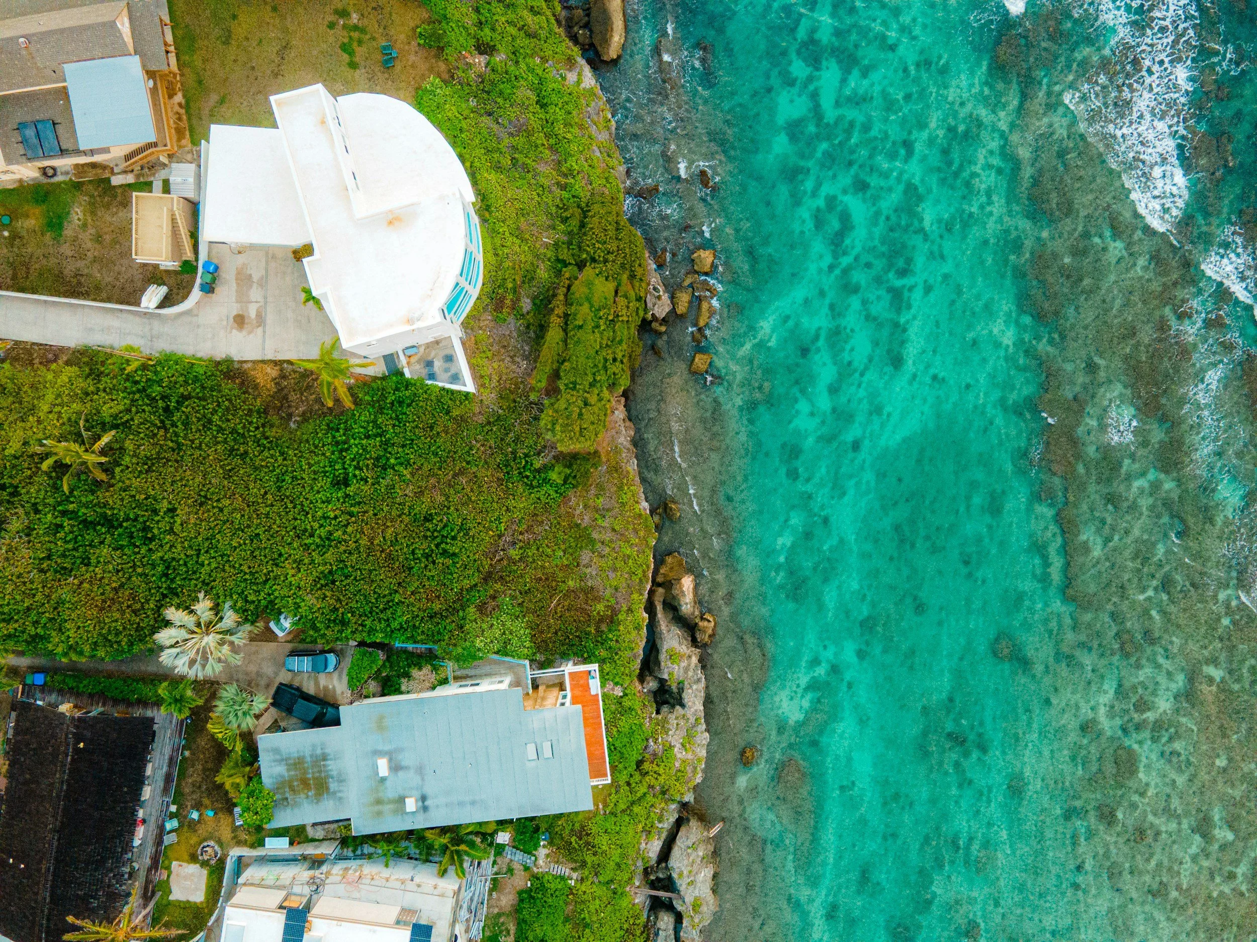 An aerial view of a coastal area with white buildings, green trees, and turquoise ocean water.