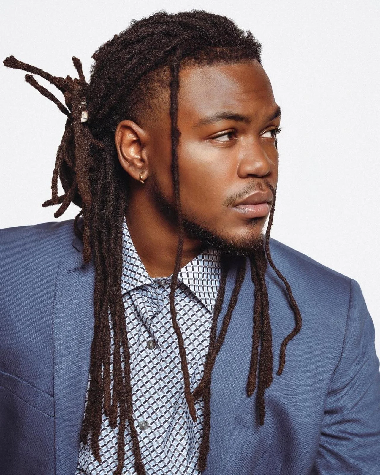 A man with long dreadlocks, wearing a navy blue suit and a patterned dress shirt, looking to the side against a plain white background.