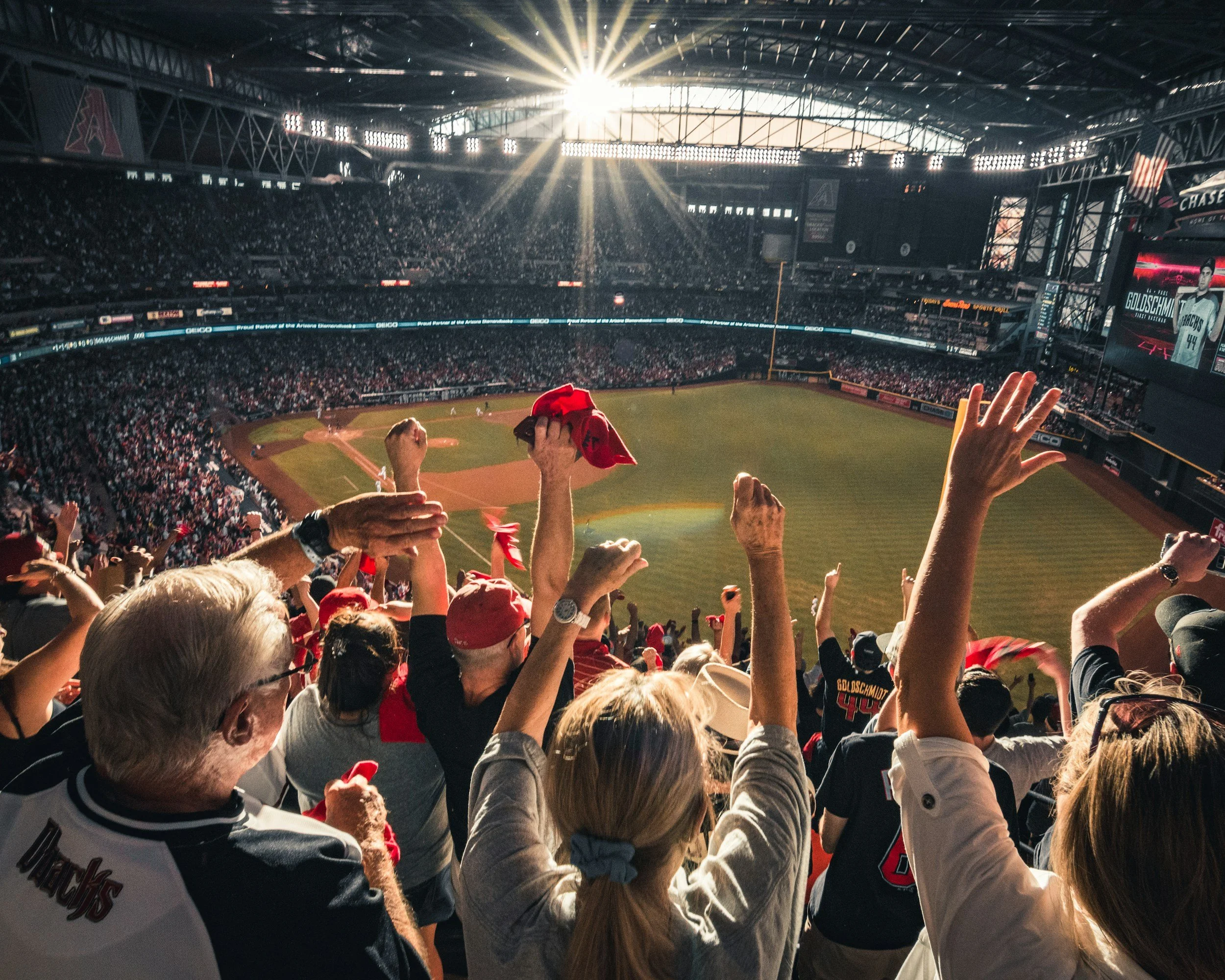 Crowd of baseball fans cheering inside a stadium with sunlight streaming through the roof and digital scoreboard displaying a player named Goldschmidt.