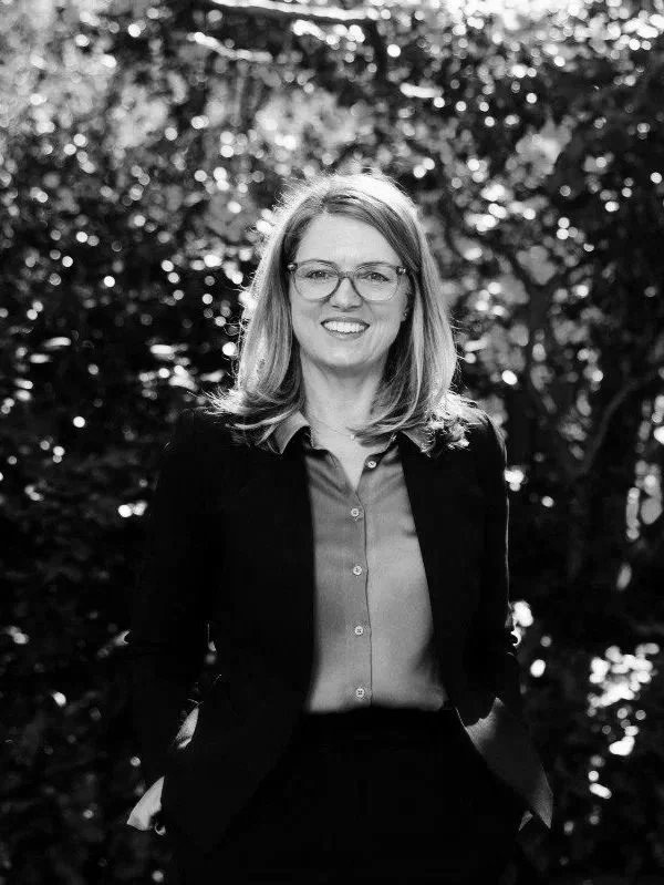 A woman with glasses, shoulder-length hair, wearing a blazer over a button-up shirt, standing outdoors with trees and sunlight in the background.