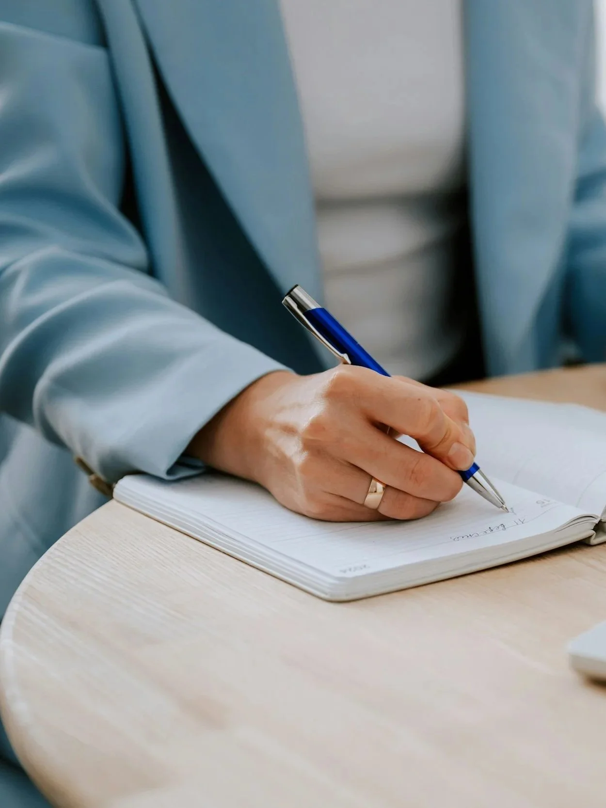 Person in a light blue blazer writing in a notebook with a blue pen, sitting at a light wooden table.