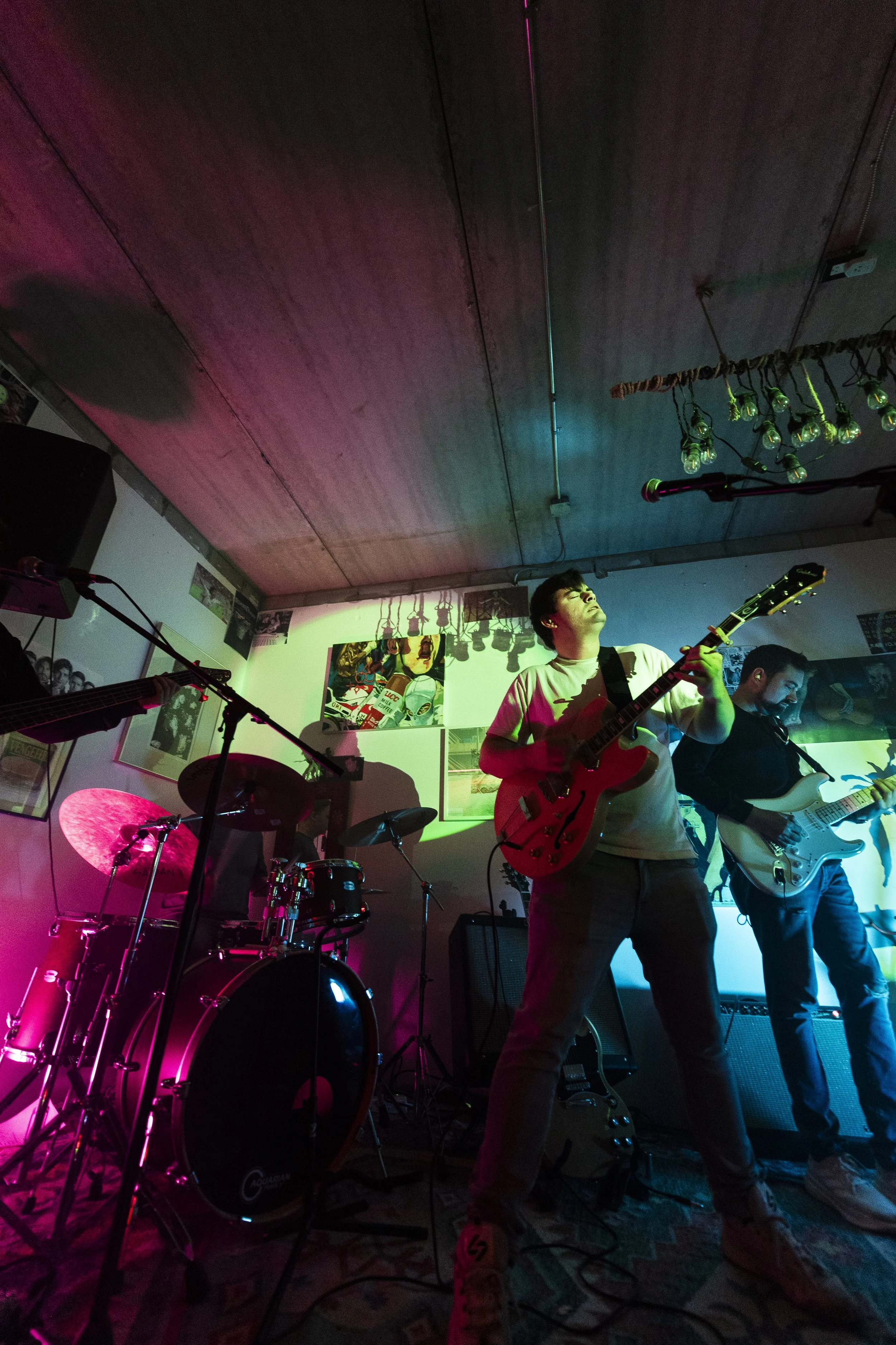 Musicians performing on stage in a dimly lit venue with colorful stage lights, including a guitarist and a drummer.