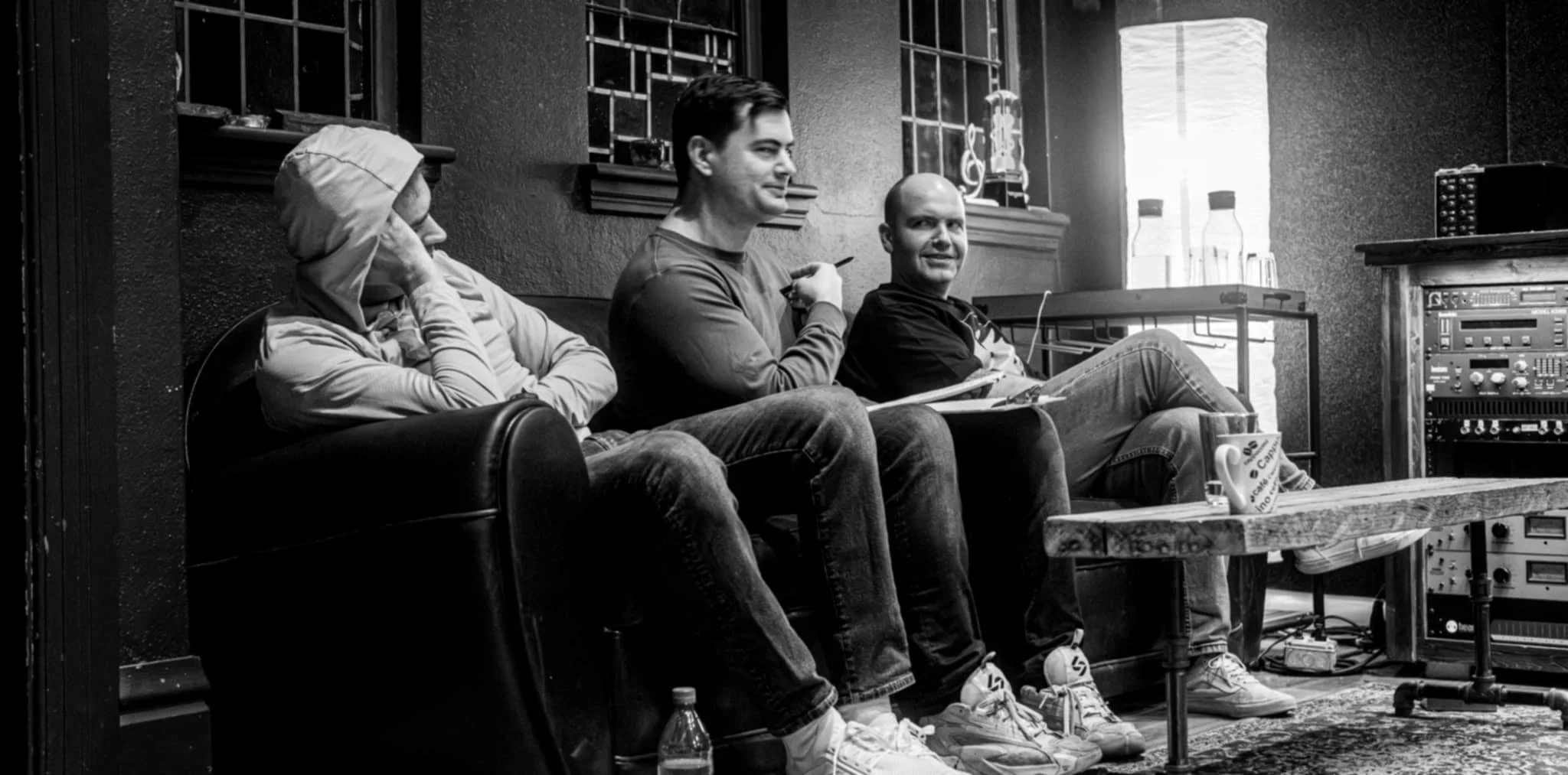 Three men sitting on a couch, with one wearing a hooded sweatshirt, in a room with sound equipment and a small table holding a mug and water bottles, black and white photo.