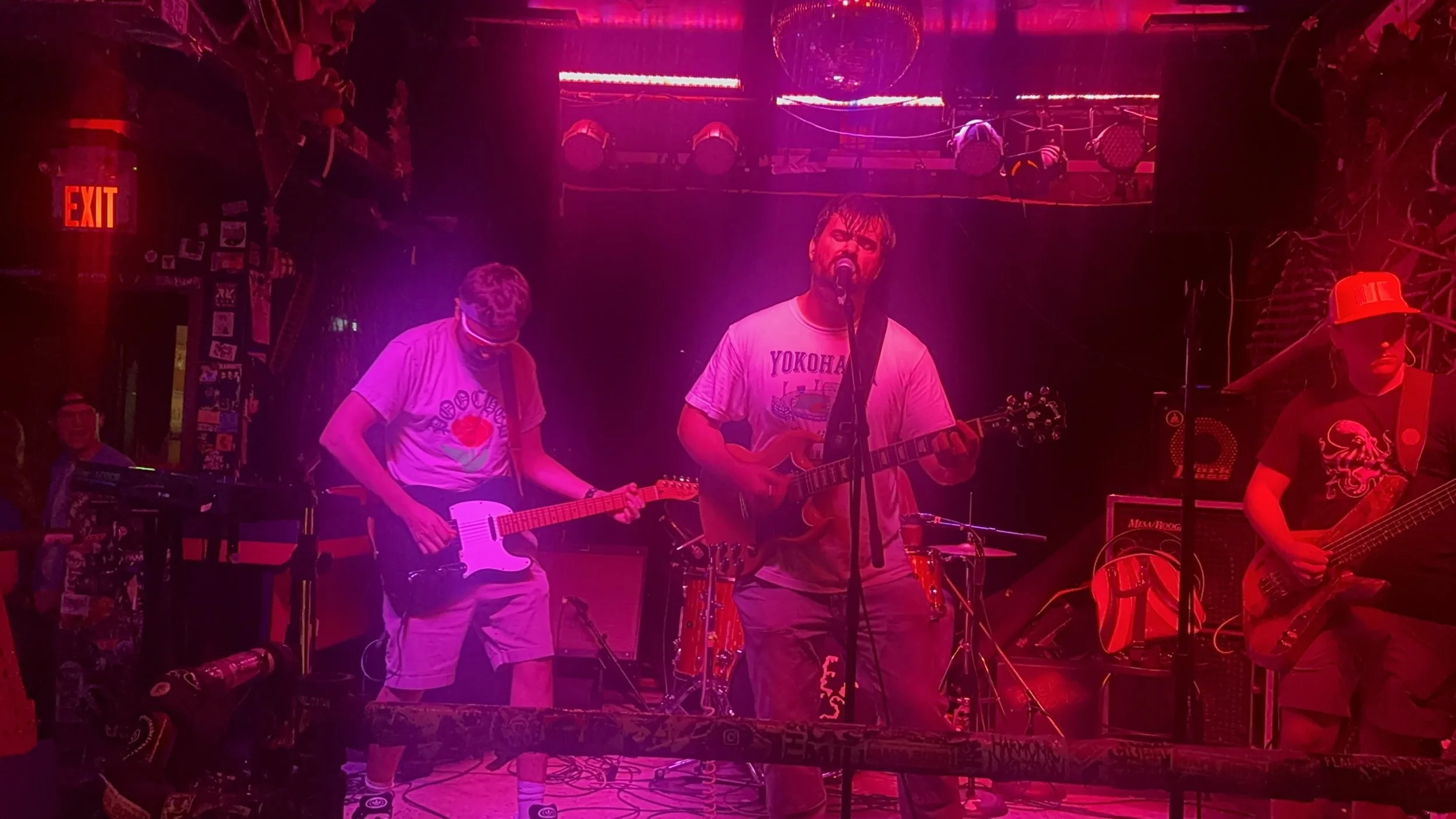 A band performs on stage with purple-pink lighting; three musicians are visible playing guitars, one singing into a microphone, in a dimly lit bar or club setting.