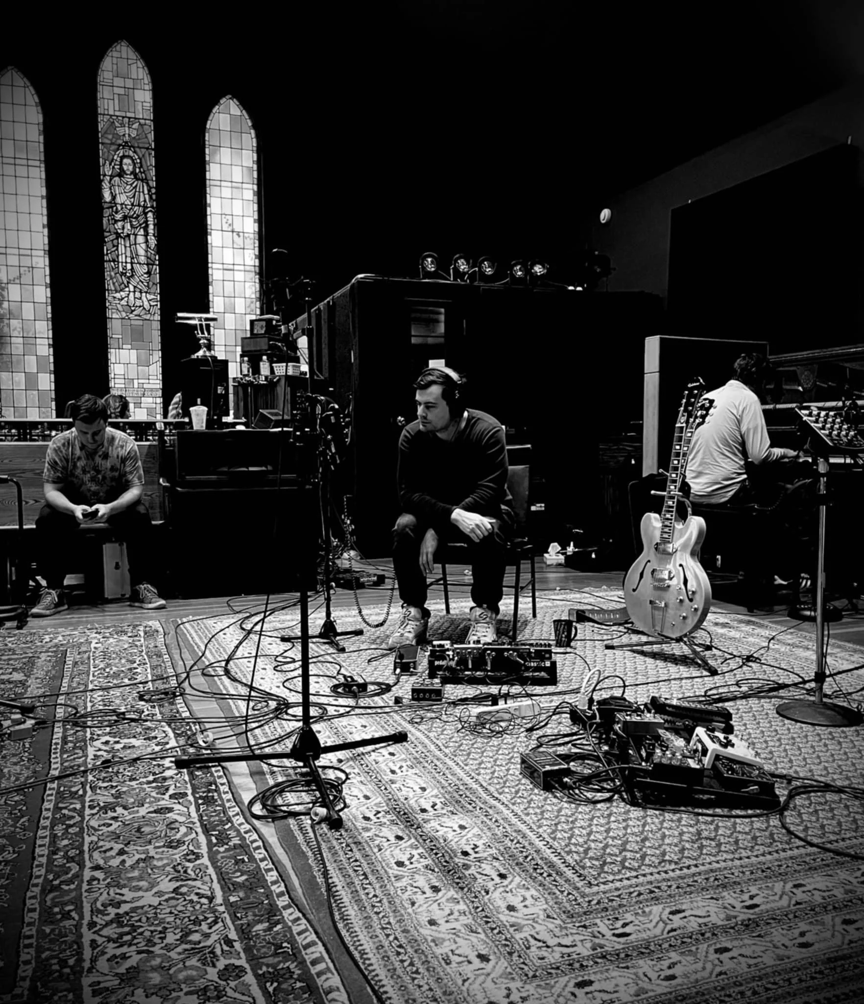 Inside view of a church with stained glass windows, a rug on the floor, and people sitting and working with music equipment, including a guitar and audio gear.