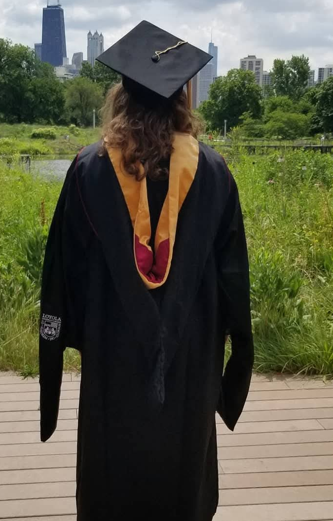 Woman wearing a black graduation gown and cap with a gold tassel and Master of Social Work hood, standing outdoors on a wooden deck with greenery and the Chicago city skyline in the background.