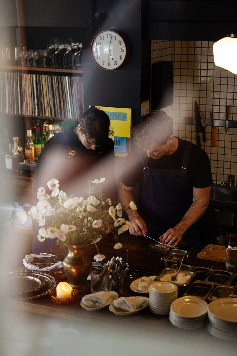 Two people cook in a dimly lit restaurant kitchen with a bouquet of white flowers and stacked plates on a counter in the foreground.