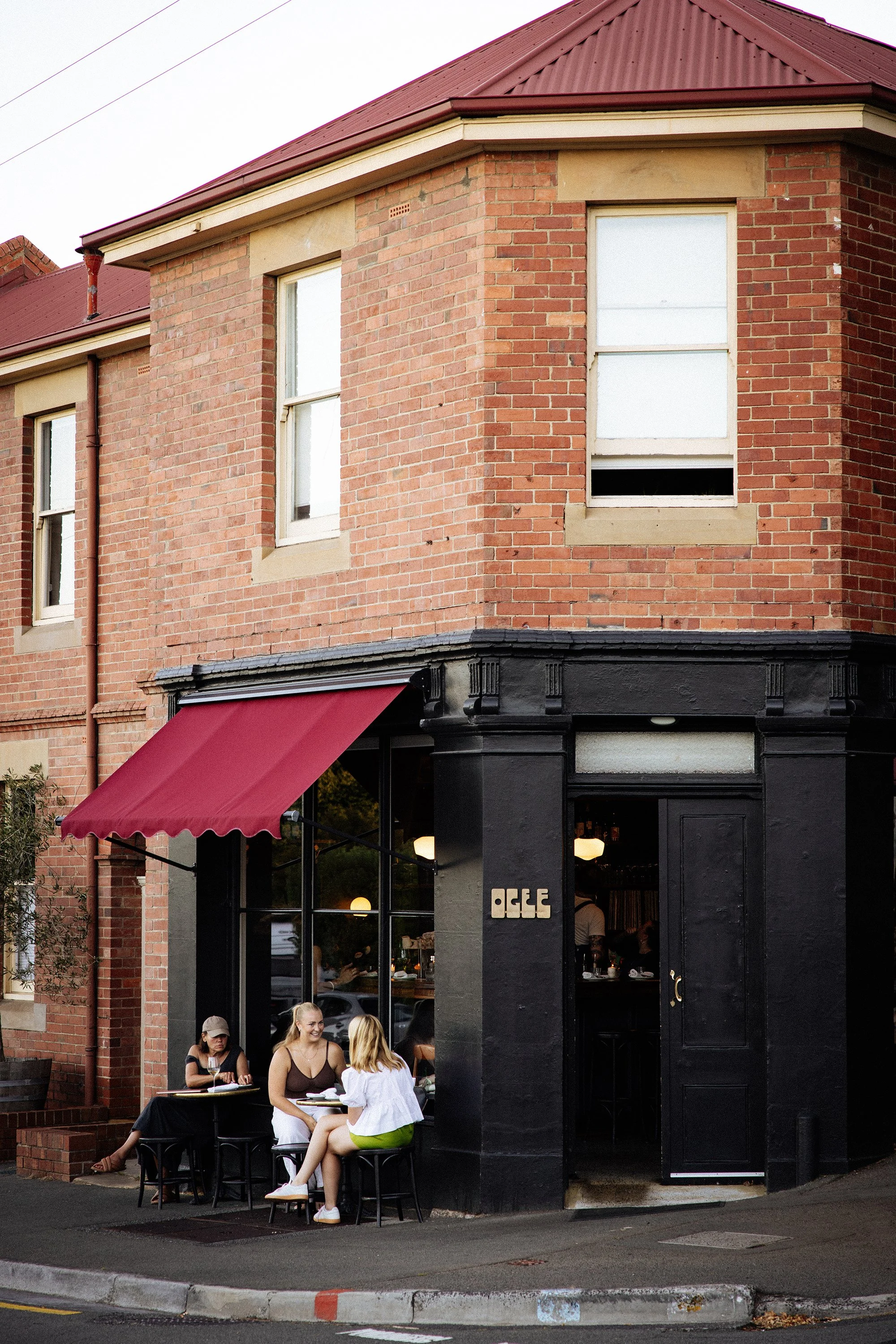 People sitting at an outdoor table in front of a restaurant with black facade and large windows, on a city street corner with brick building above, during daytime.