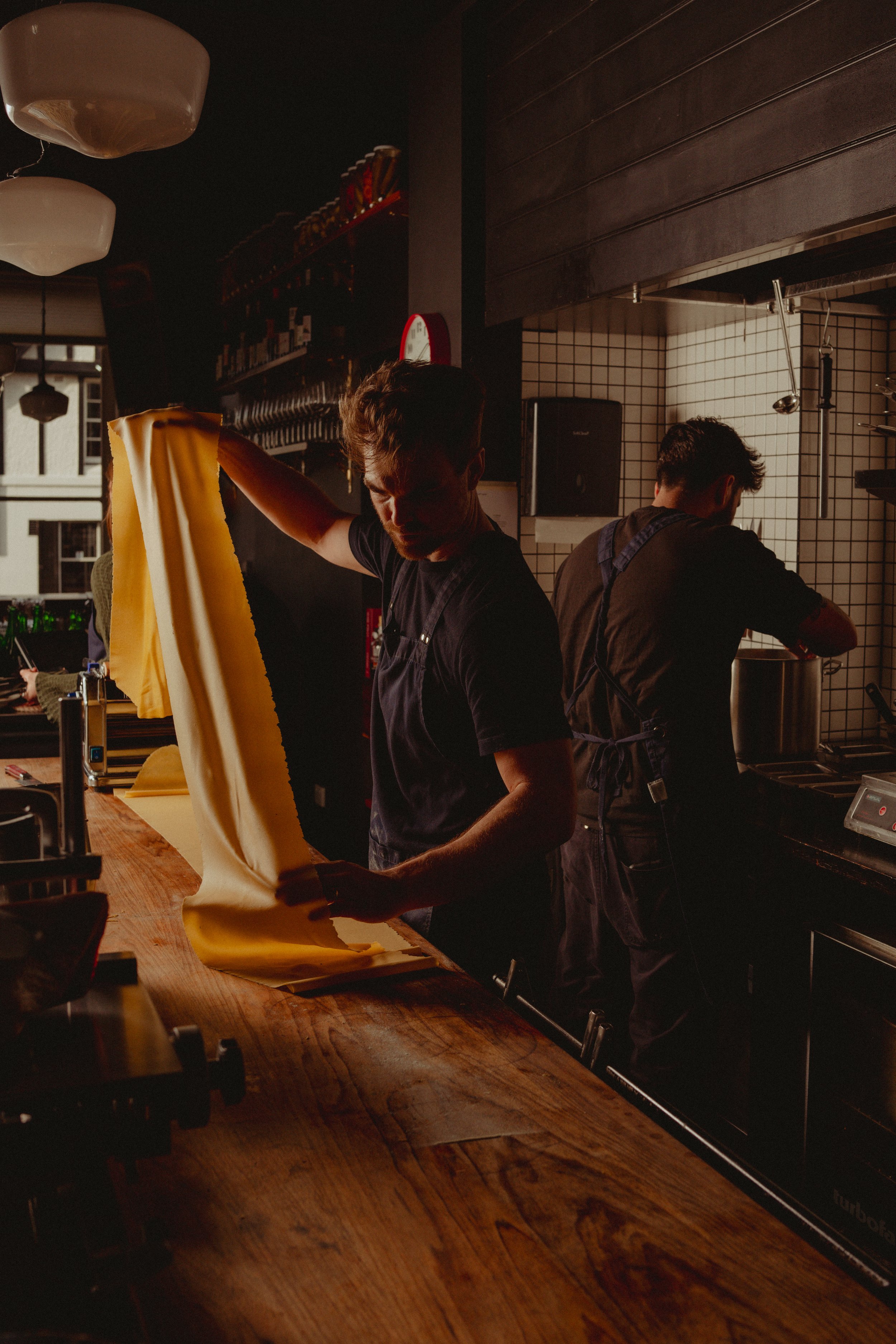 Two men working in a restaurant kitchen with one folding a large sheet of dough and the other stirring a pot on the stove. The kitchen has white tiled walls and dark wooden cabinets.