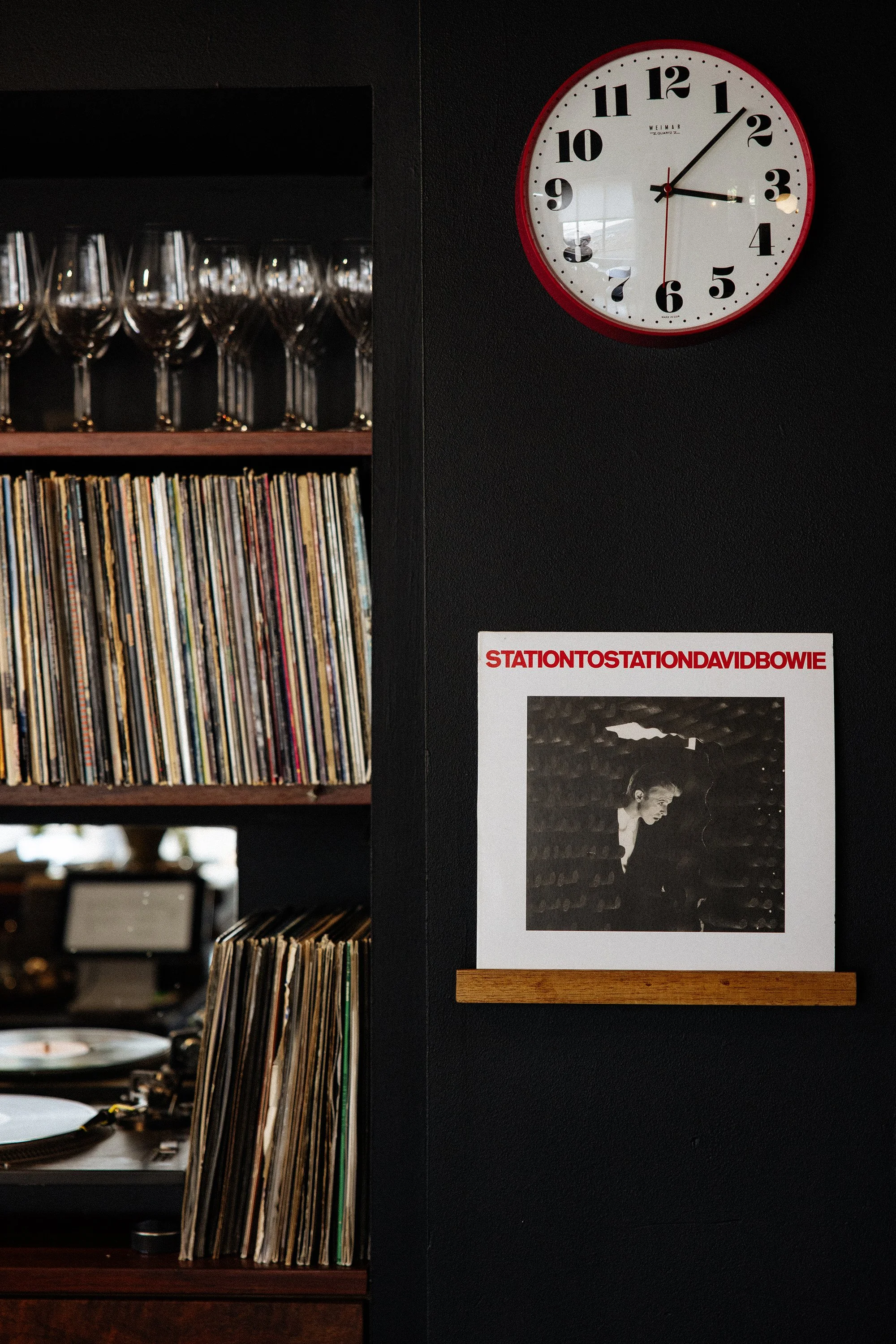 An interior scene with a wall clock showing the time at 2:15, a shelf filled with vinyl records, and wine glasses on another shelf. There is a poster of David Bowie with the text "STATIONTOSTATION DAVID BOWIE" beneath it.