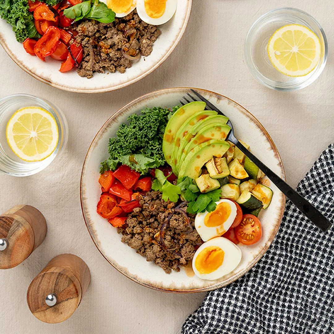 Beef mince breakfast bowls with sliced avocado, roasted courgette and capsicum, fresh kale, parsley, cherry tomatoes and boiled eggs, served on ceramic plates.