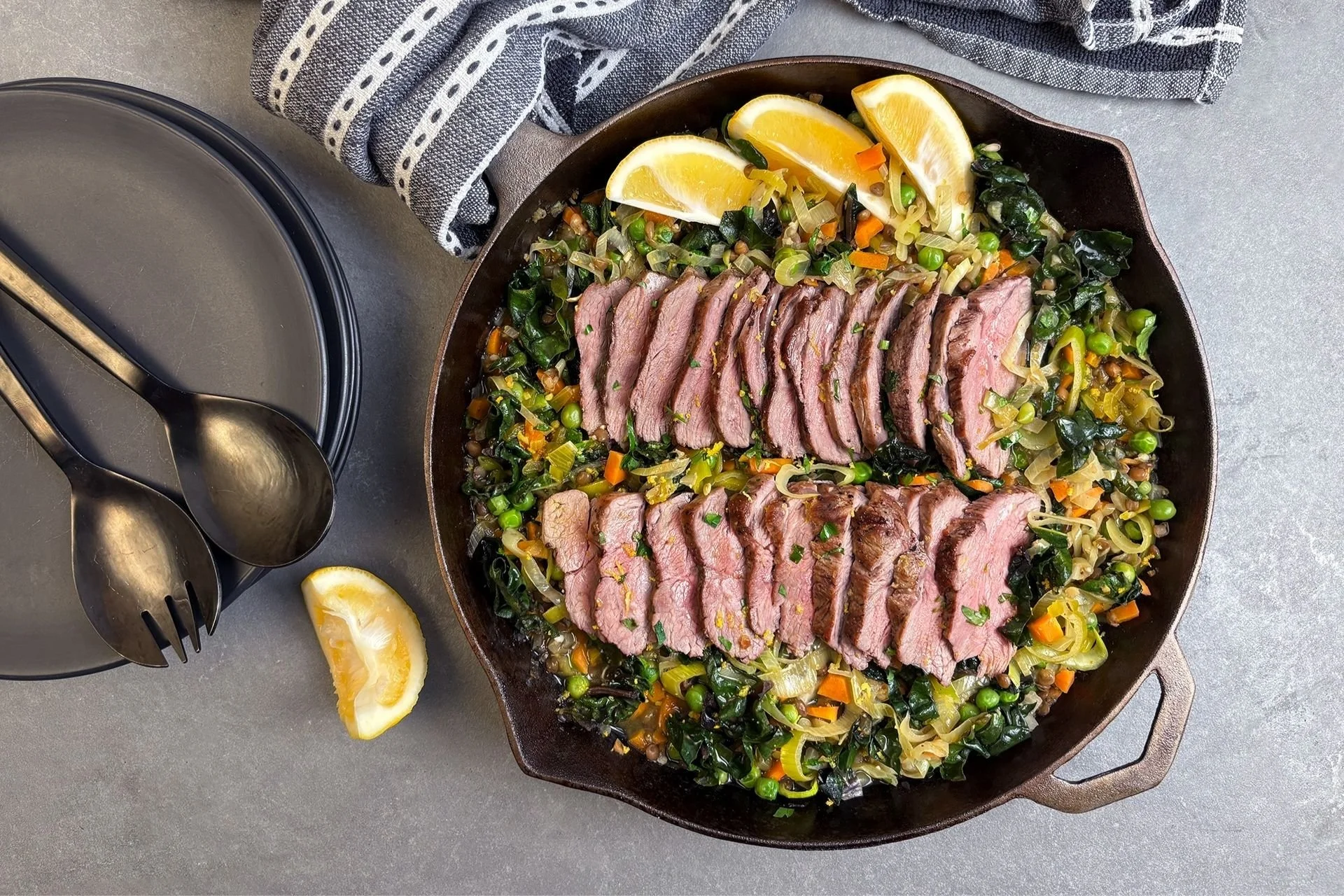 Sliced lamb rump served over sautéed greens, peas, leeks and diced carrots in a skillet, garnished with fresh herbs and lemon wedges.