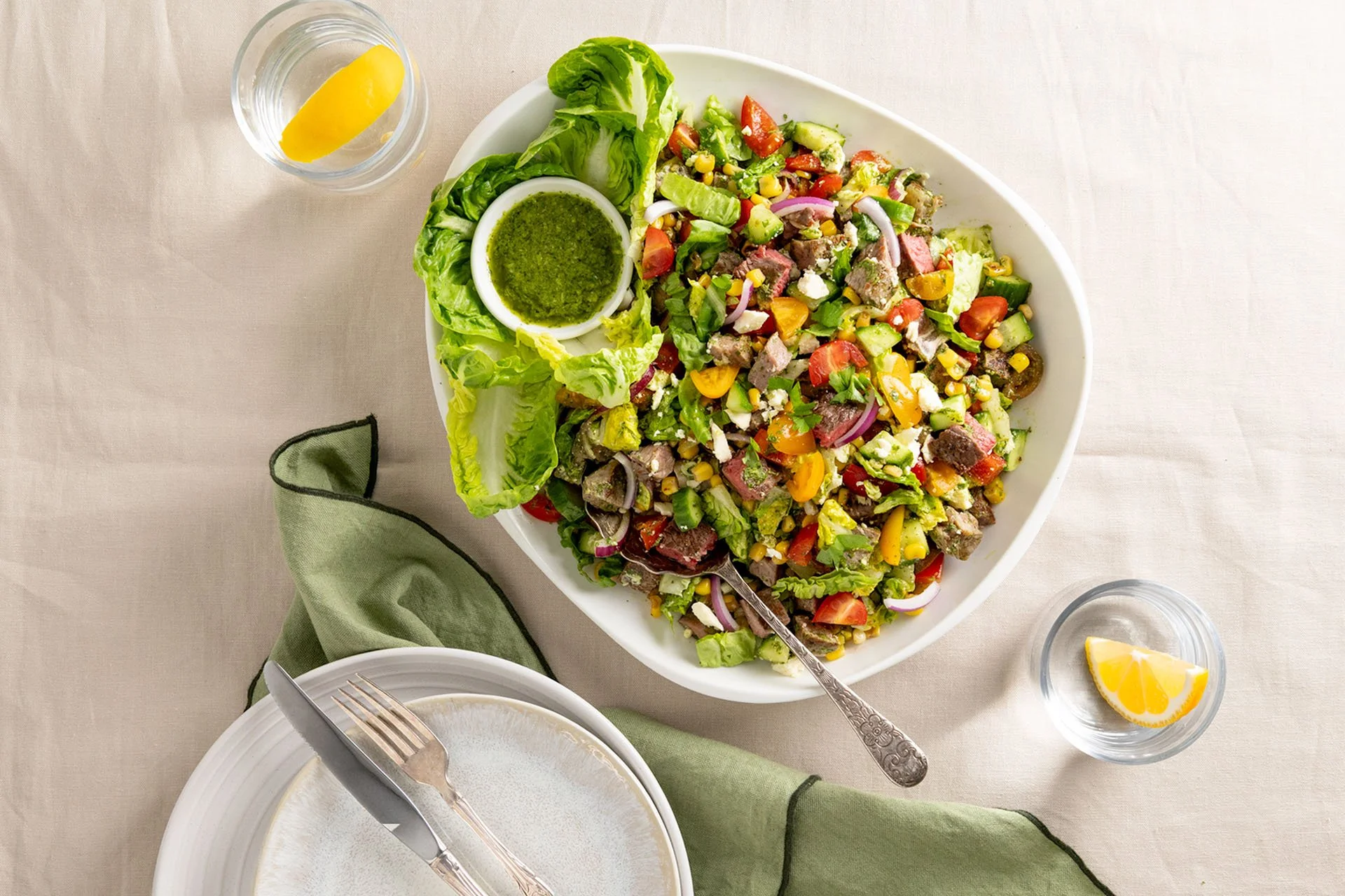 Chopped steak salad with fresh tomatoes, corn, cucumber, red onion, feta and green lettuce, served with a bowl of green chimichurri dressing and lemon wedges.