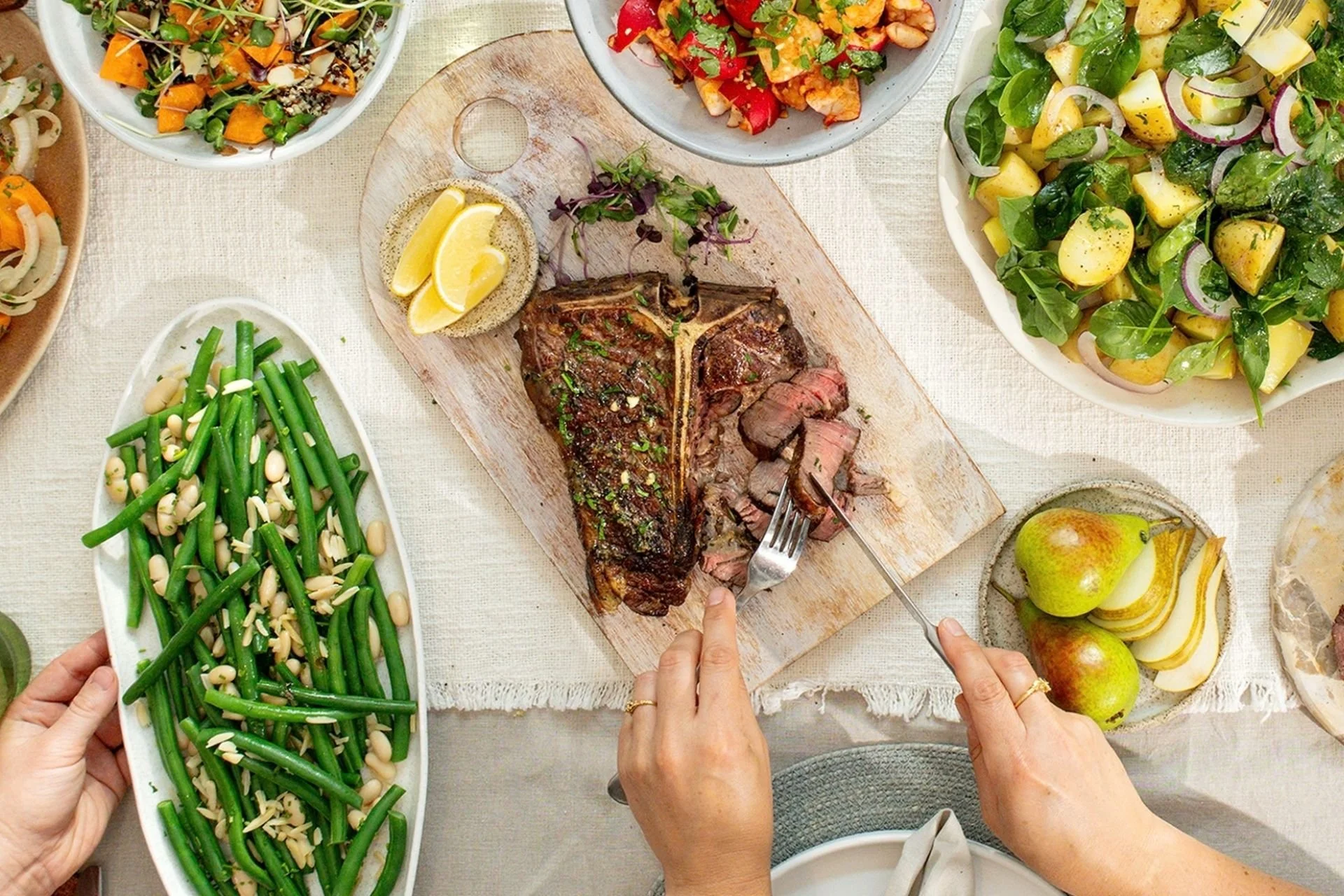 Hands slicing a steak at a shared table with green beans and salads