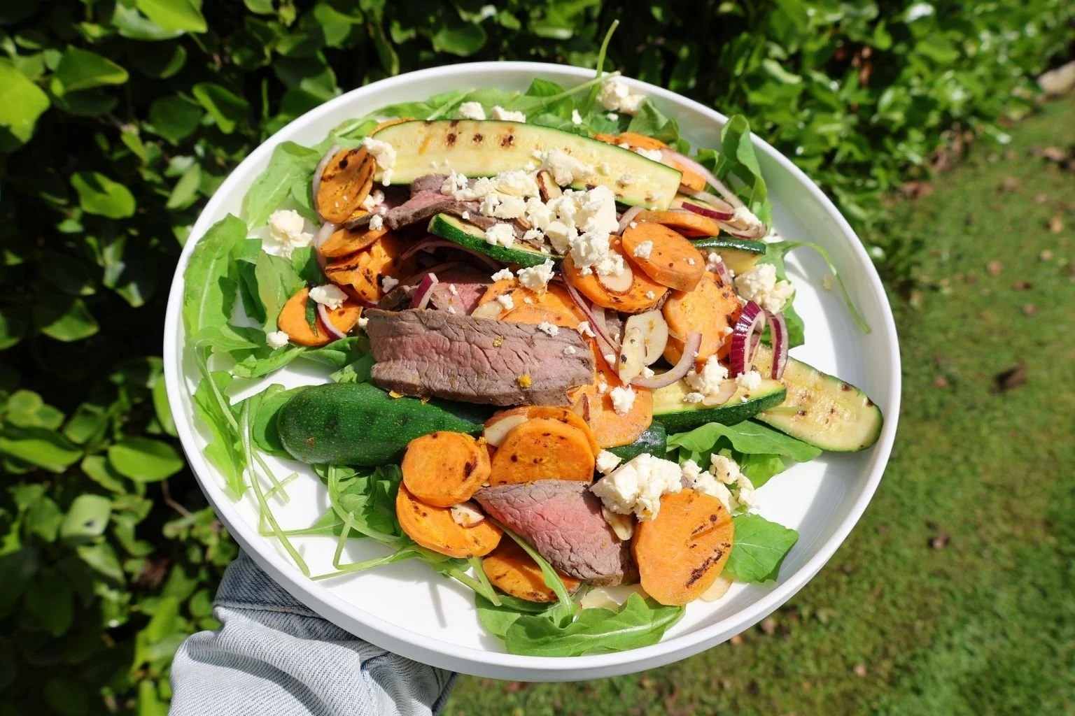 Grilled steak, kumara and courgette salad with fresh rocket, red onion and crumbled feta, served on a white plate outdoors.
