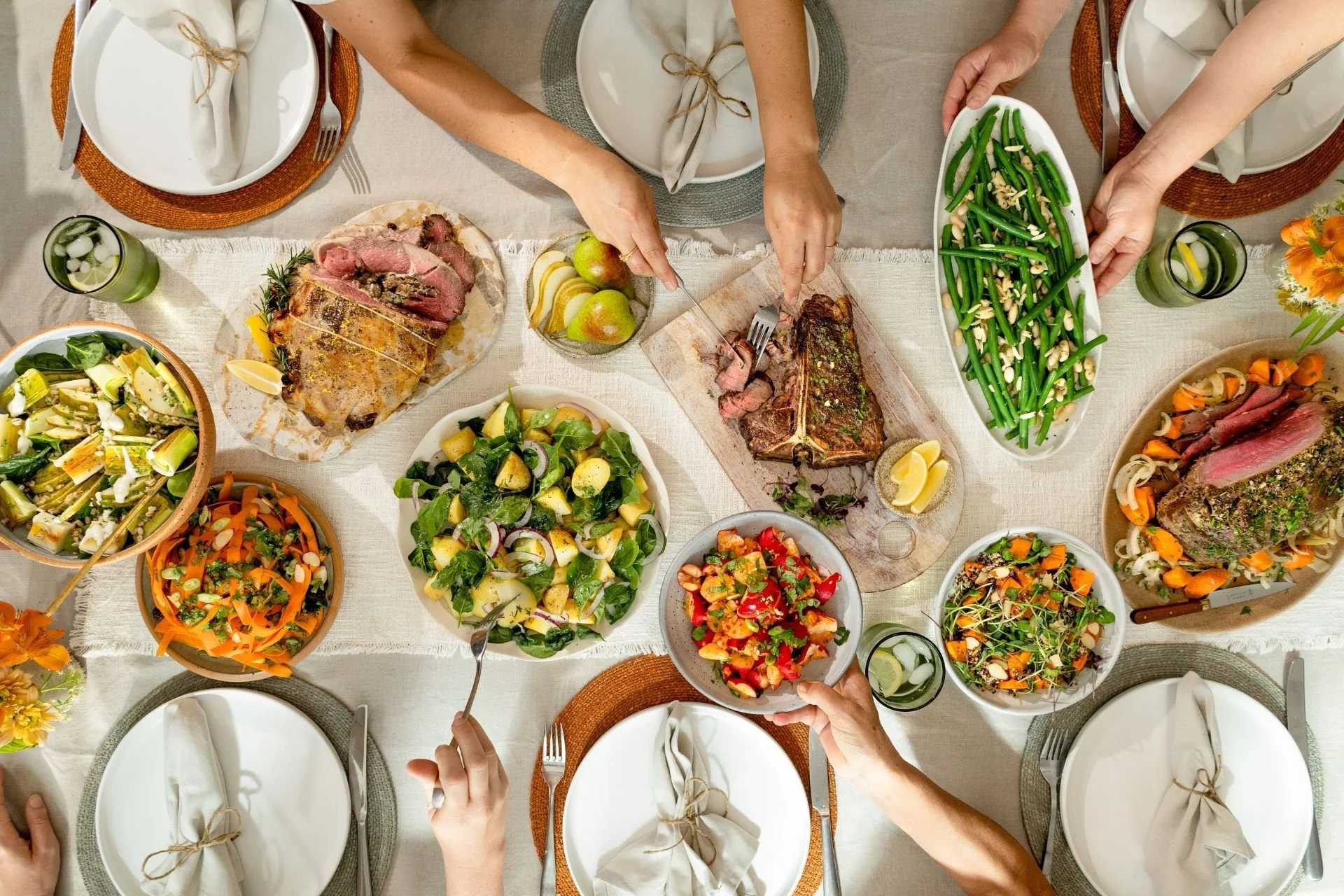 Overhead view of a shared family table with roast lamb, T-bone steak, roast beef, green beans, potato salad and fresh vegetable salads.