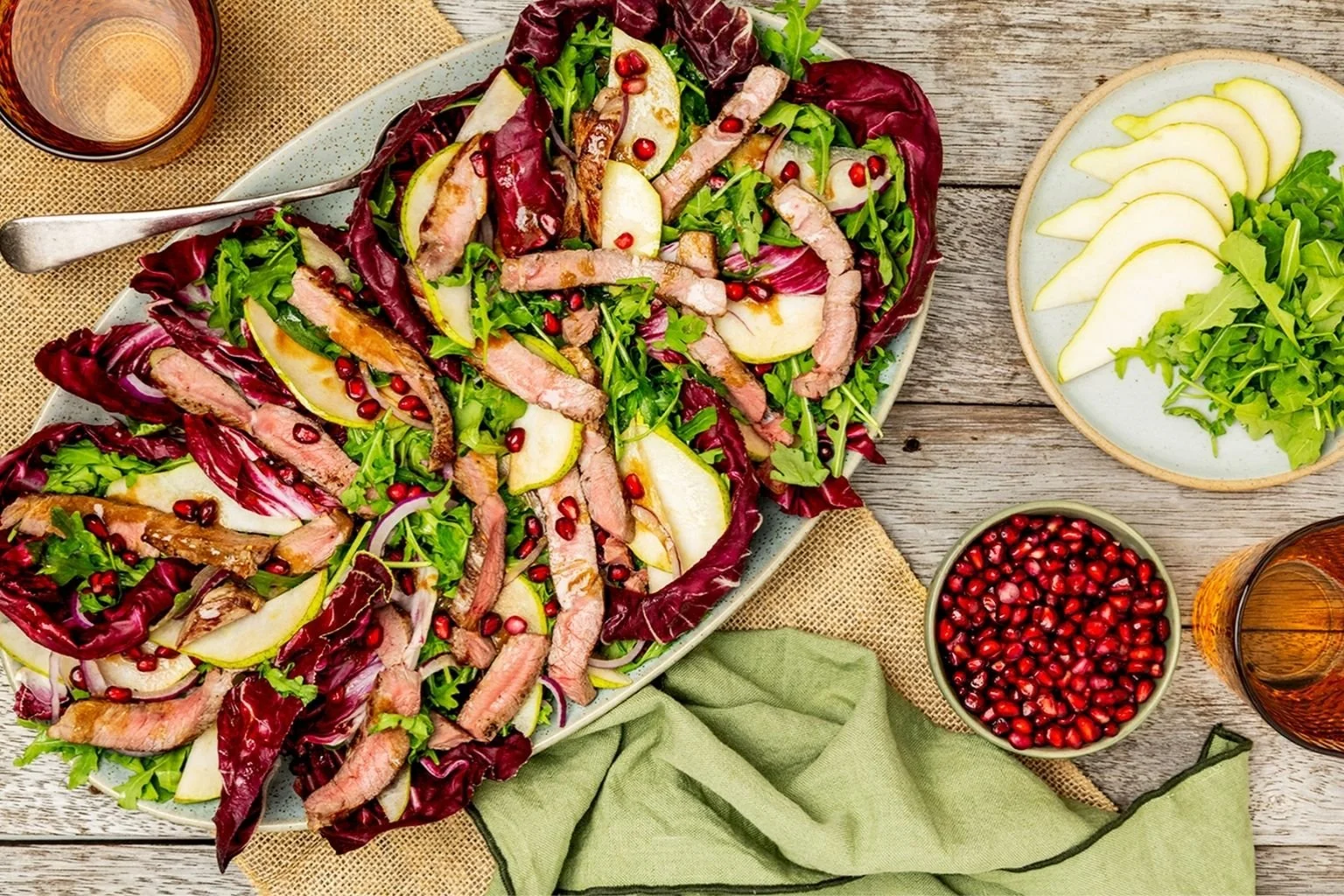 Sliced grilled steak salad with pear, pomegranate seeds, rocket, radicchio and red onion, served on a platter with extra pear slices and pomegranate seeds on the side.