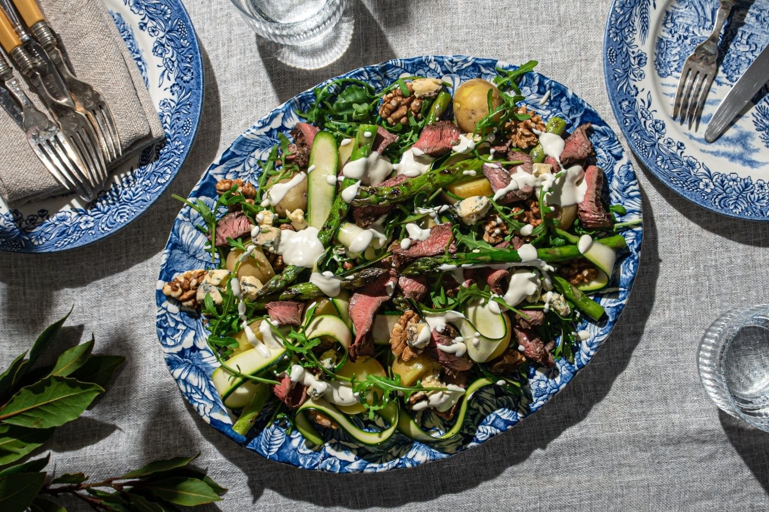 Sliced grilled beef rump steak salad with asparagus, courgette ribbons, baby potatoes, rocket, walnuts and blue cheese, drizzled with creamy dressing on a blue and white platter.
