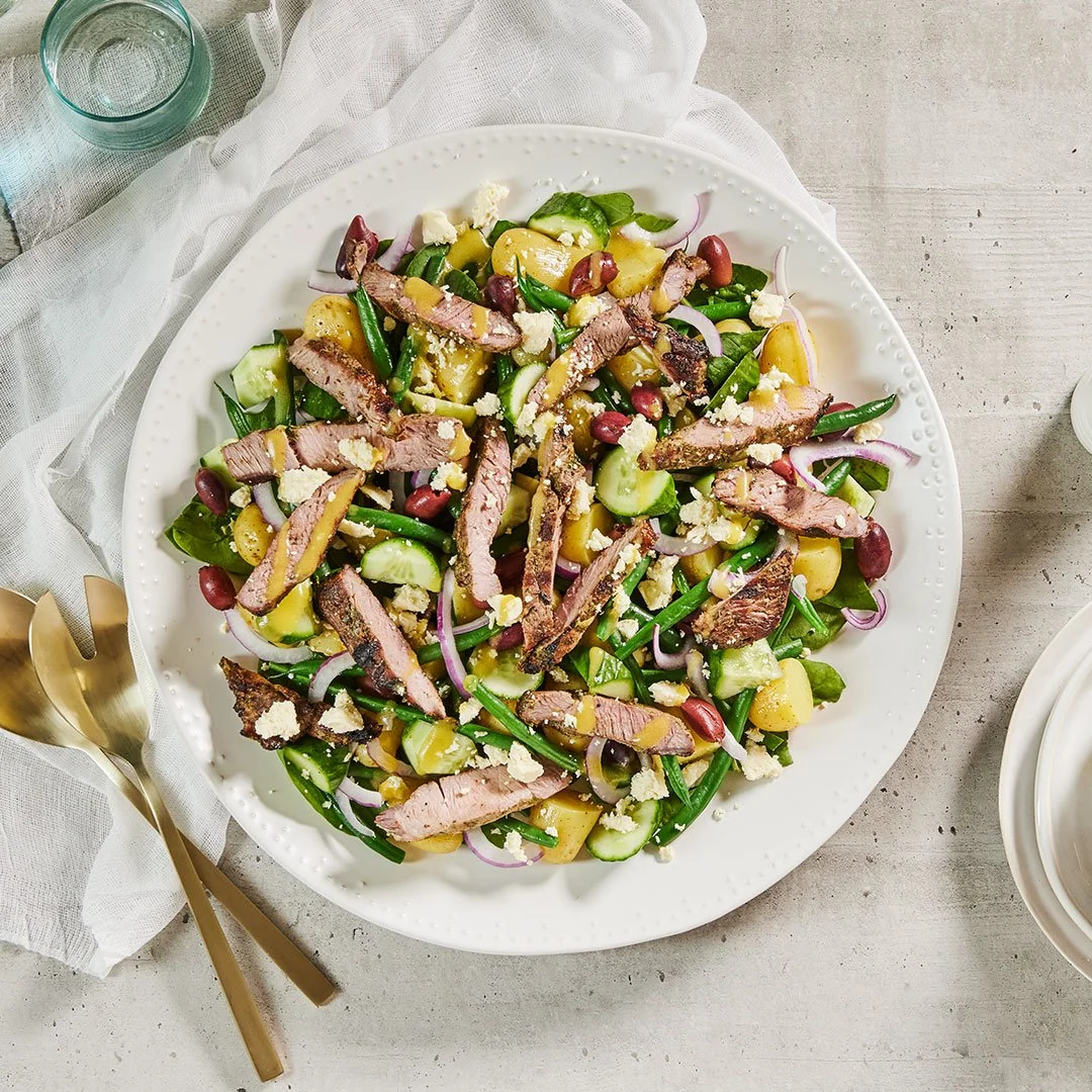 Greek lamb salad with sliced grilled lamb, baby potatoes, green beans, cucumber, red onion, spinach, kidney beans and crumbled feta on a white serving platter.
