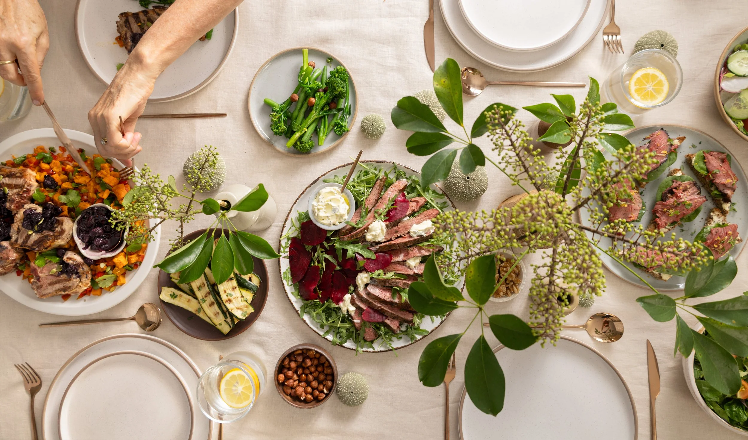Overhead view of a shared dining table featuring sliced beef steak salad with beetroot and creamy dressing, grilled lamb chops, roasted vegetable salad, grilled courgettes and green vegetables.