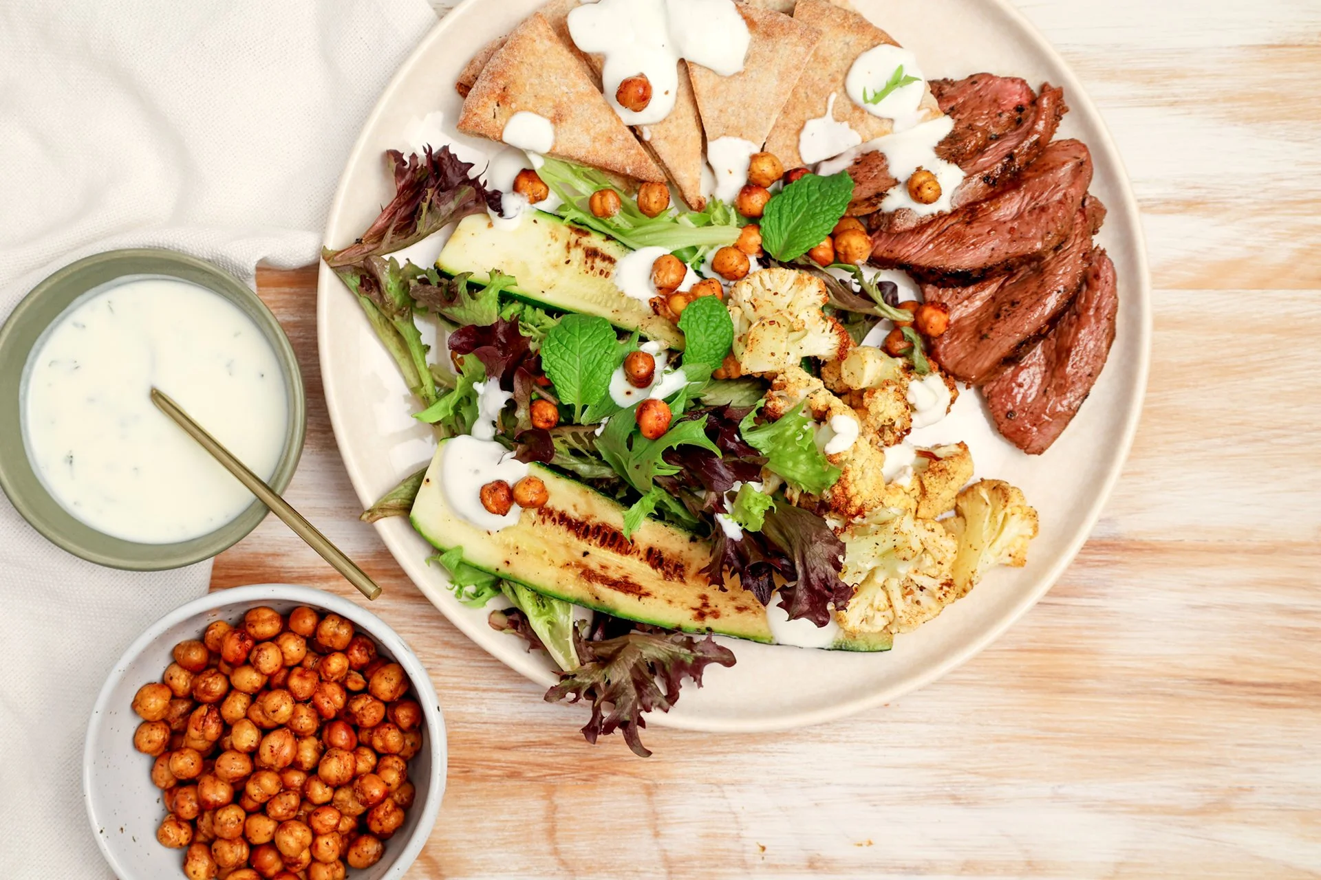 Moroccan-spiced lamb rump sliced and served with grilled courgette, roasted cauliflower, crispy chickpeas, mixed salad leaves and flatbread, drizzled with creamy yoghurt dressing and garnished with fresh mint.