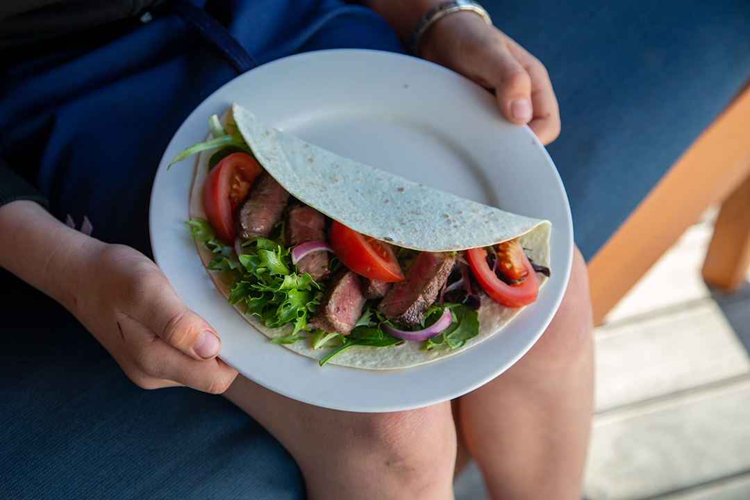 Simple beef steak wrap with sliced steak, tomato, red onion and fresh salad greens, held on a white plate outdoors.