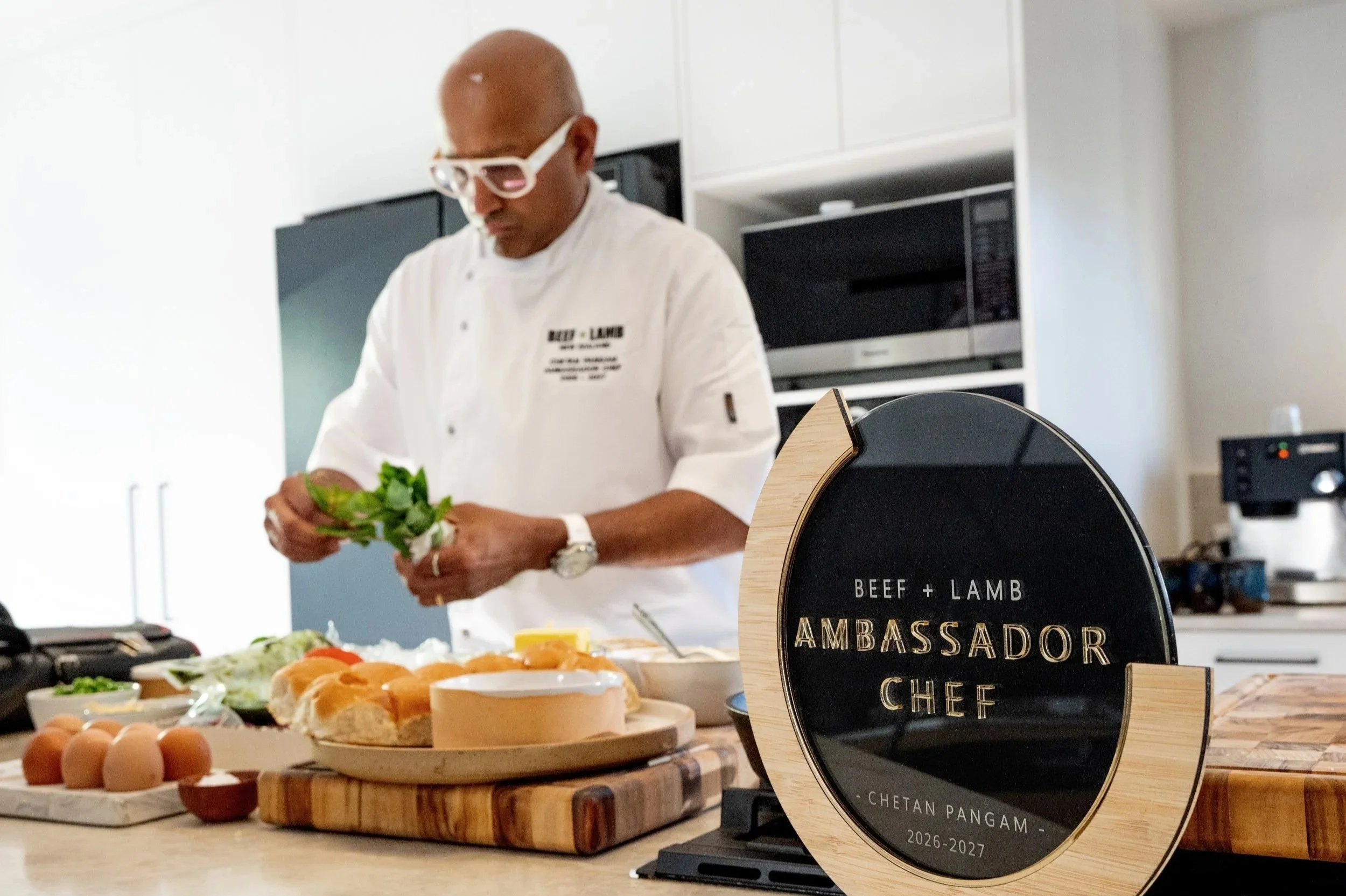 chef in a white chefs jacket with bald head and white glasses makign food in a domestic kitchen with his Ambassador Chef plaque in foreground.
