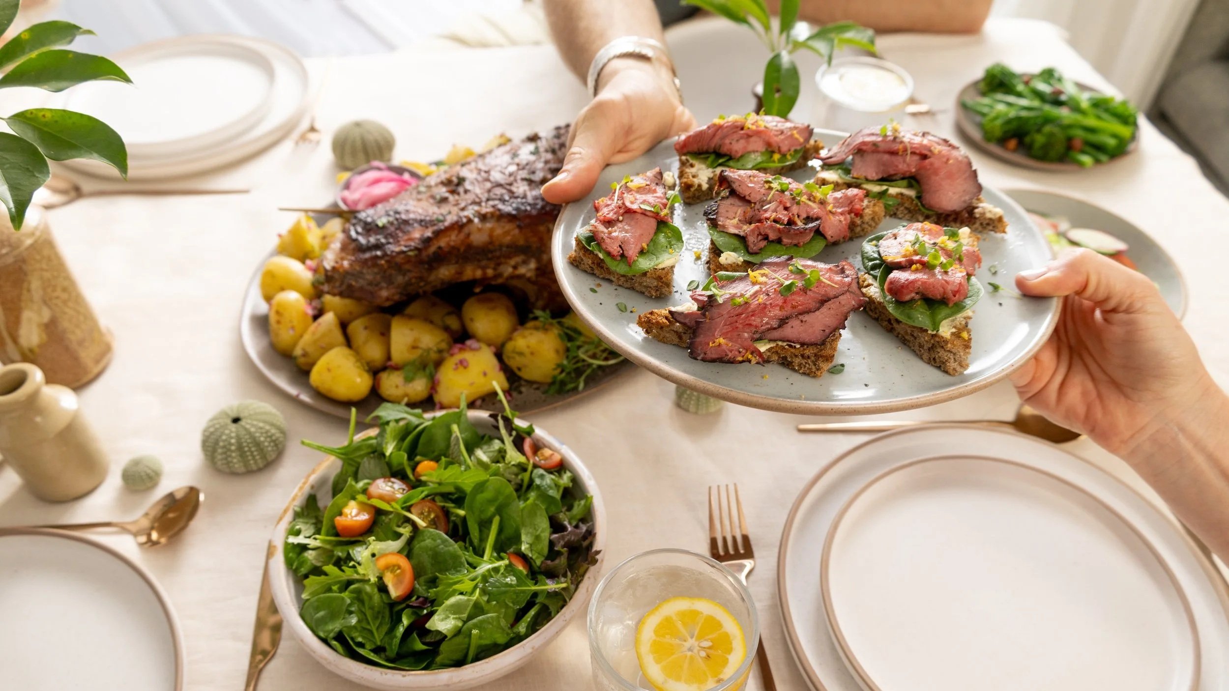 Hands passing a plate of rare roast beef on toasted bread, with a roast, potatoes and salad shared across a table
