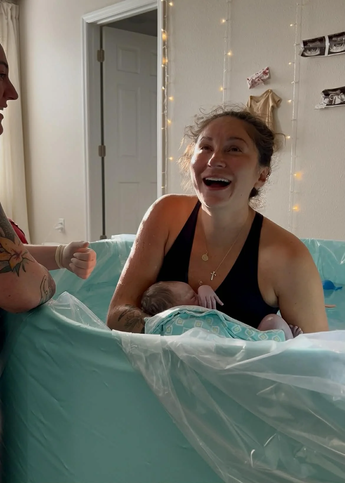 A woman is laughing during her water birth, holding her newborn baby, with a man nearby in a tub. The room has string lights and photos on the wall.