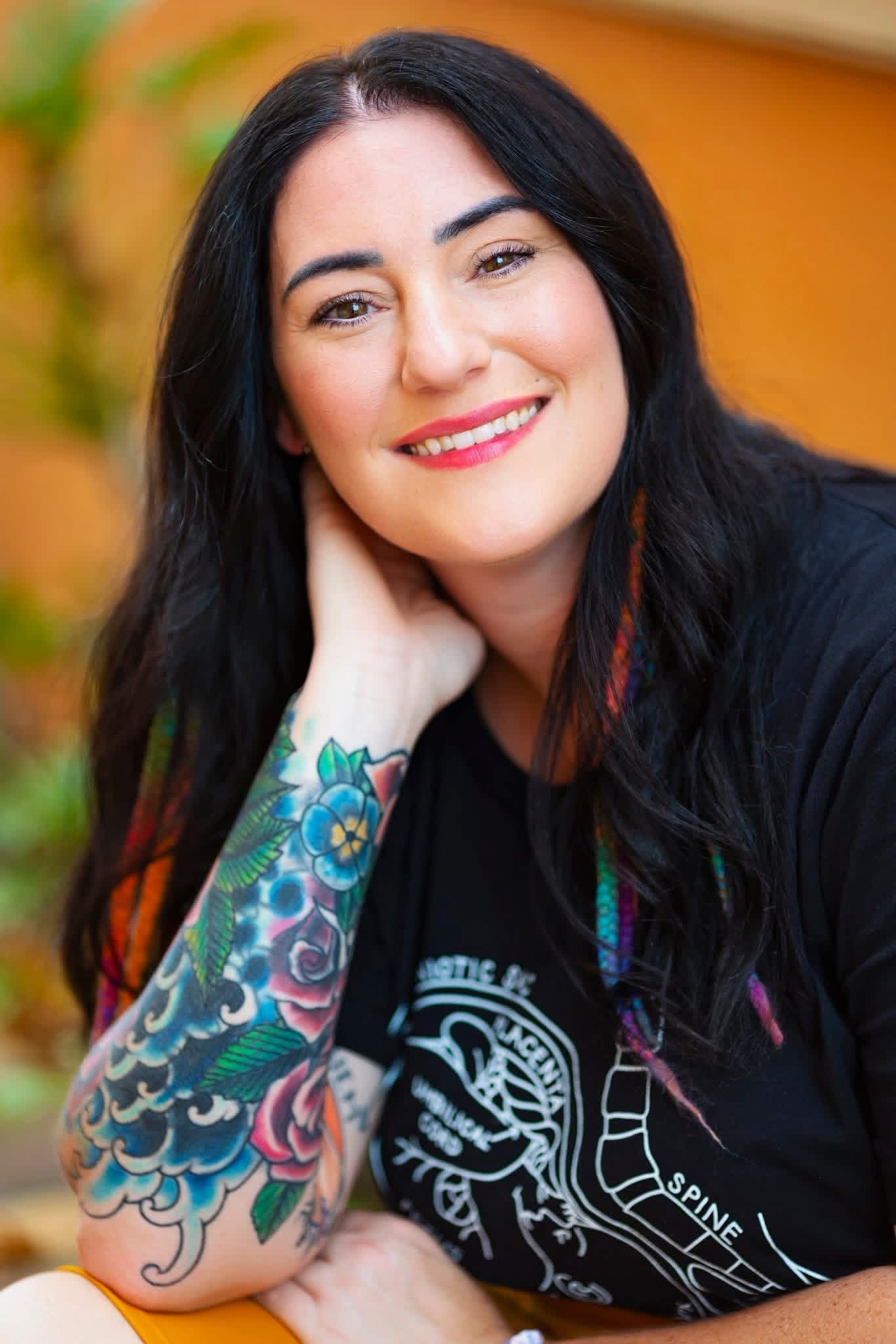 A woman with long black hair and light makeup, smiling with her hand resting on her neck, has colorful floral tattoos on her left arm, is wearing a black T-shirt with white text and graphics, and is outdoors with blurred autumn foliage in the background.